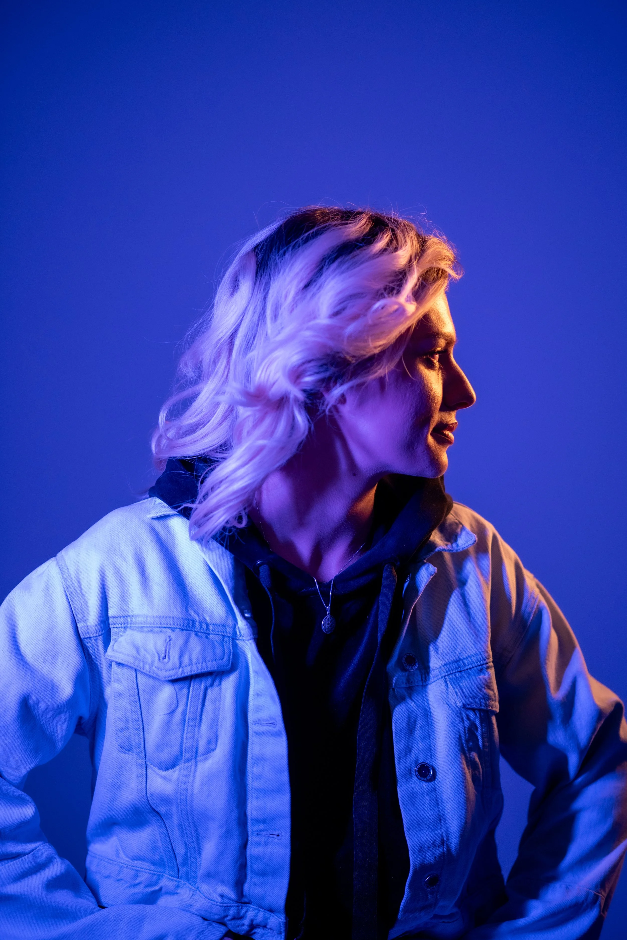 Profile of a woman with blonde hair, wearing a jacket and a hoodie, illuminated with purple and blue lighting.
