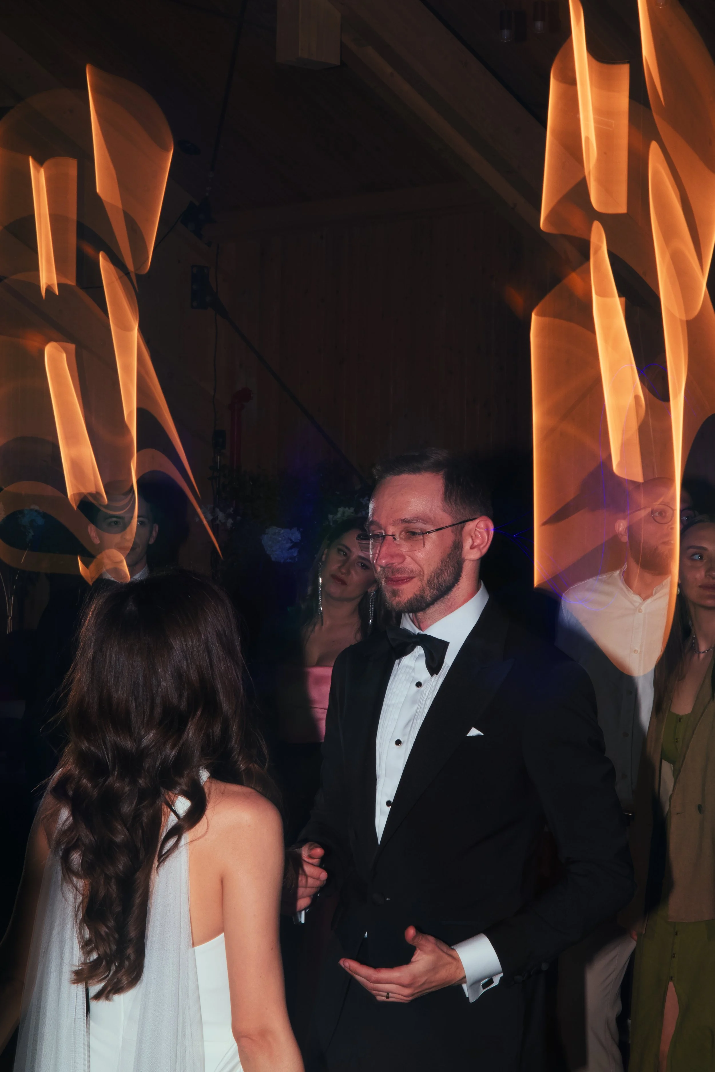 A man in a tuxedo and bow tie talking to a woman with long dark hair at a formal event, with others in the background and orange streaks of light in motion above.