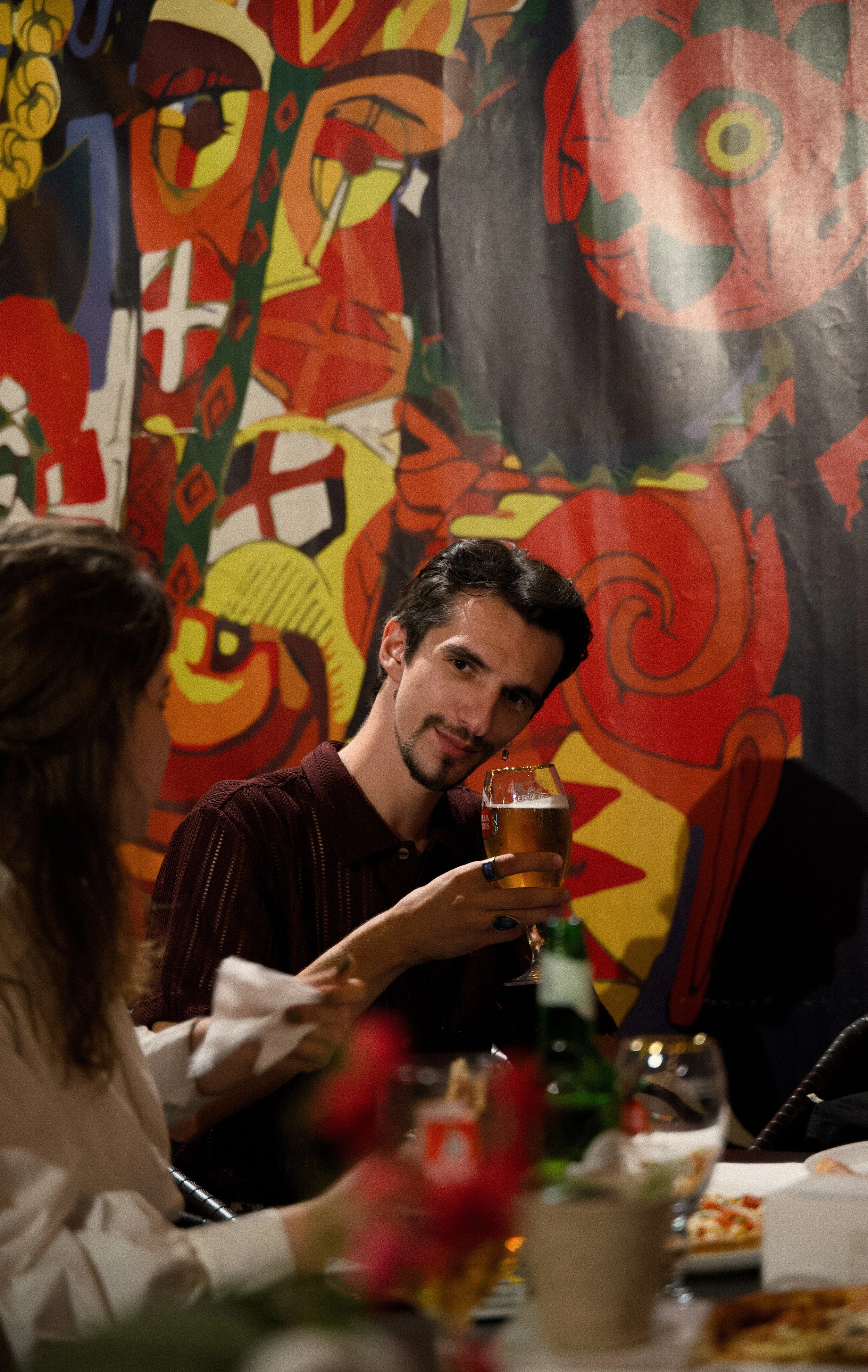 Man holding a glass of beer at a dinner gathering, sitting at a table with food and drinks, with colorful abstract artwork on the wall in the background.