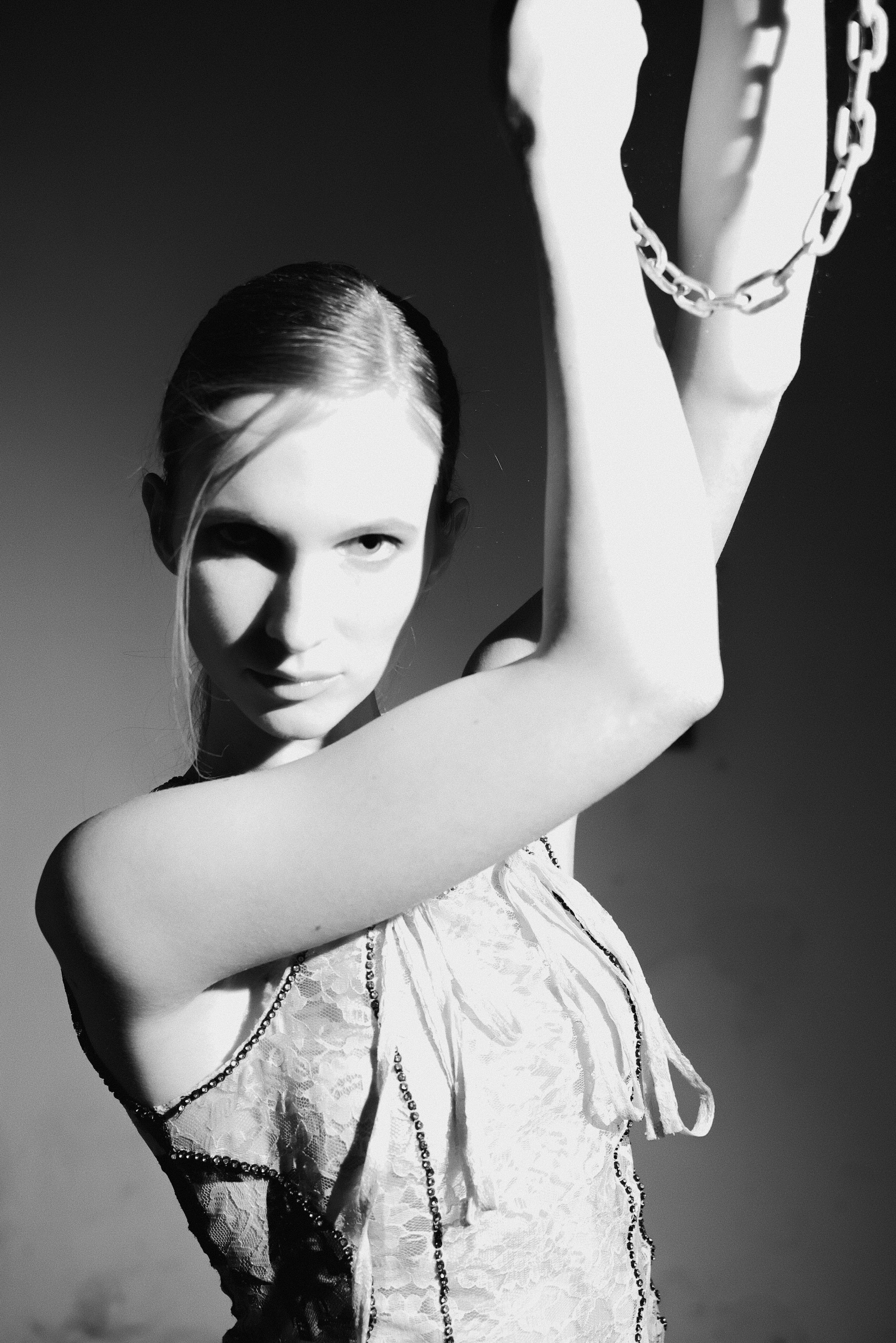A black and white photo of a woman with wet hair, wearing a lace lingerie top, holding a chain near her shoulder, with intense eye contact and an assertive expression.