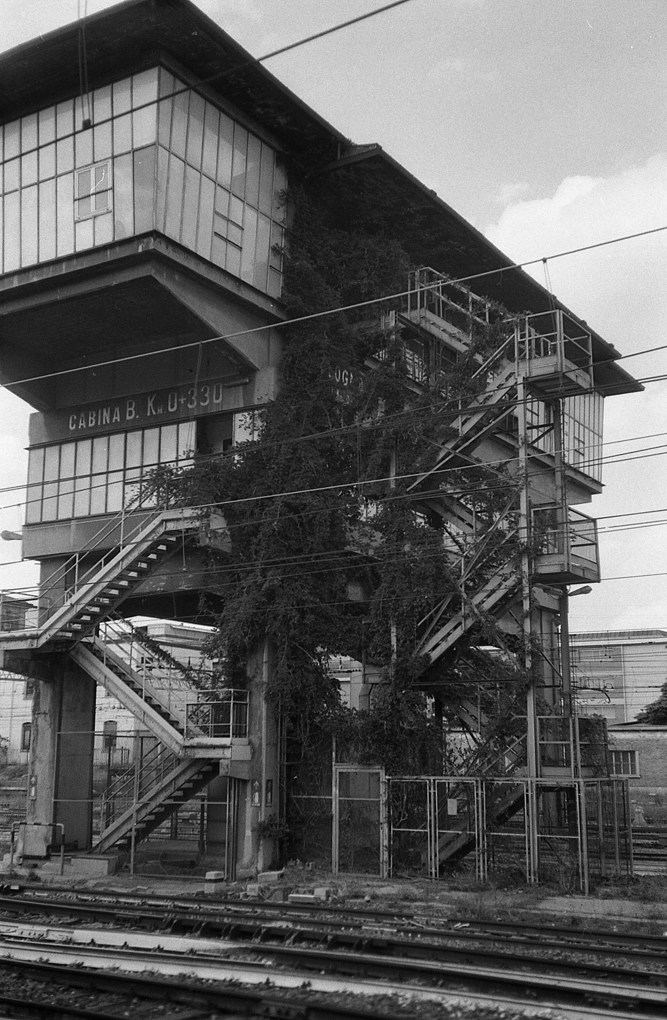 Black and white photo of an overgrown train station control tower with stairs and multiple levels, adjacent to railway tracks.