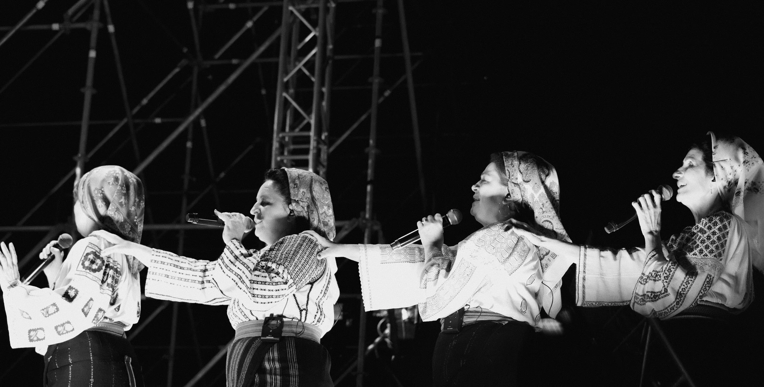 Four women dressed in traditional embroidered garments, wearing headscarves, performing on stage with microphones, in front of a dark background and metal scaffolding.