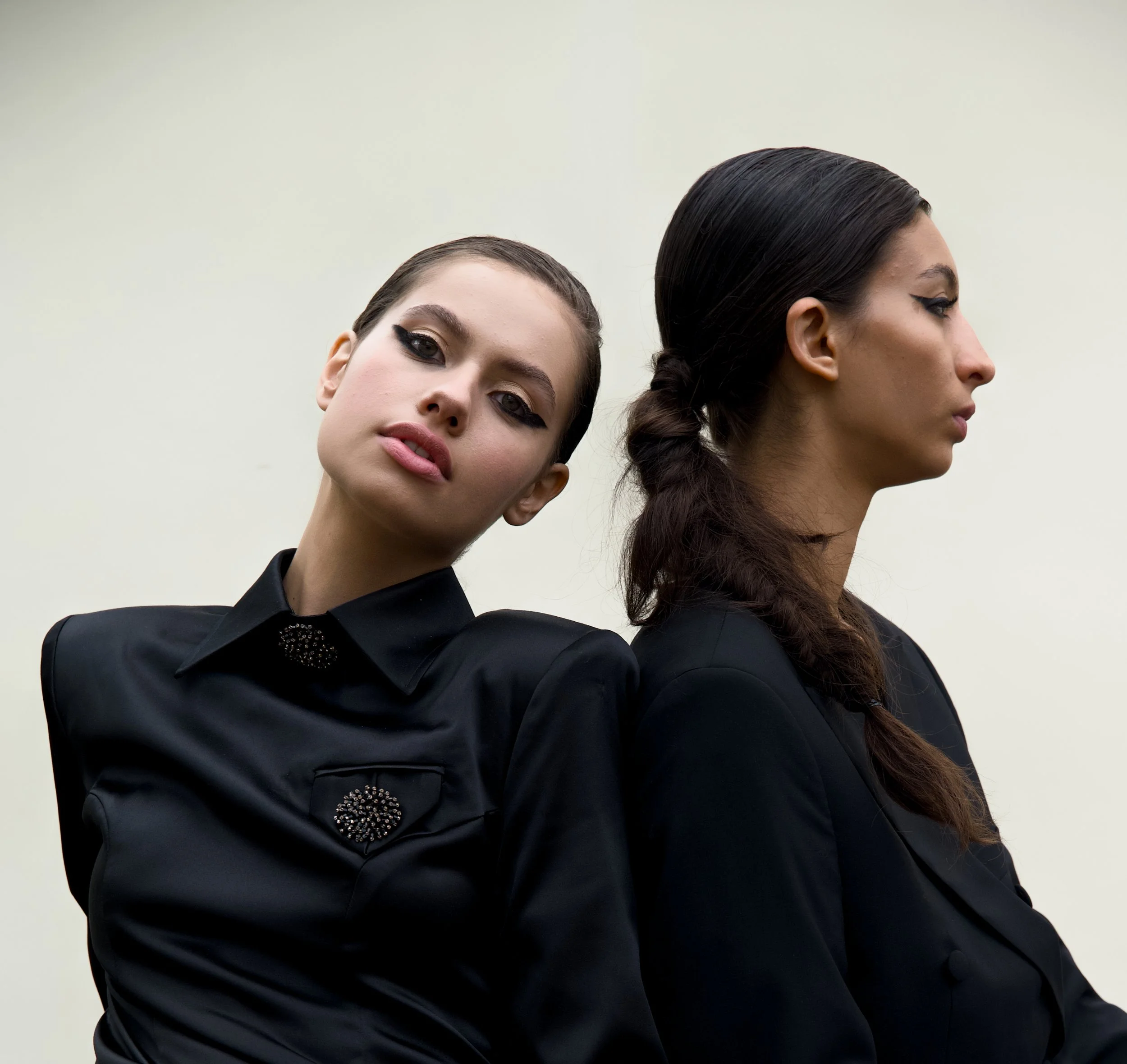 Two women with dark hair and makeup, wearing black clothing, standing against a plain light-colored background. One woman faces forward, tilting her head and gazing directly, while the other is in profile with her eyes closed.