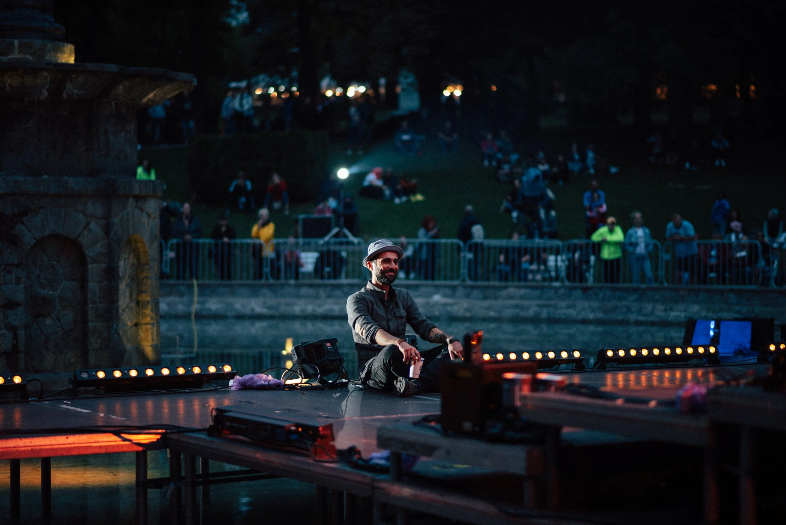 A DJ smiling and sitting cross-legged on a stage at an outdoor event during dusk, with a crowd of people sitting and standing behind a fence in the background.