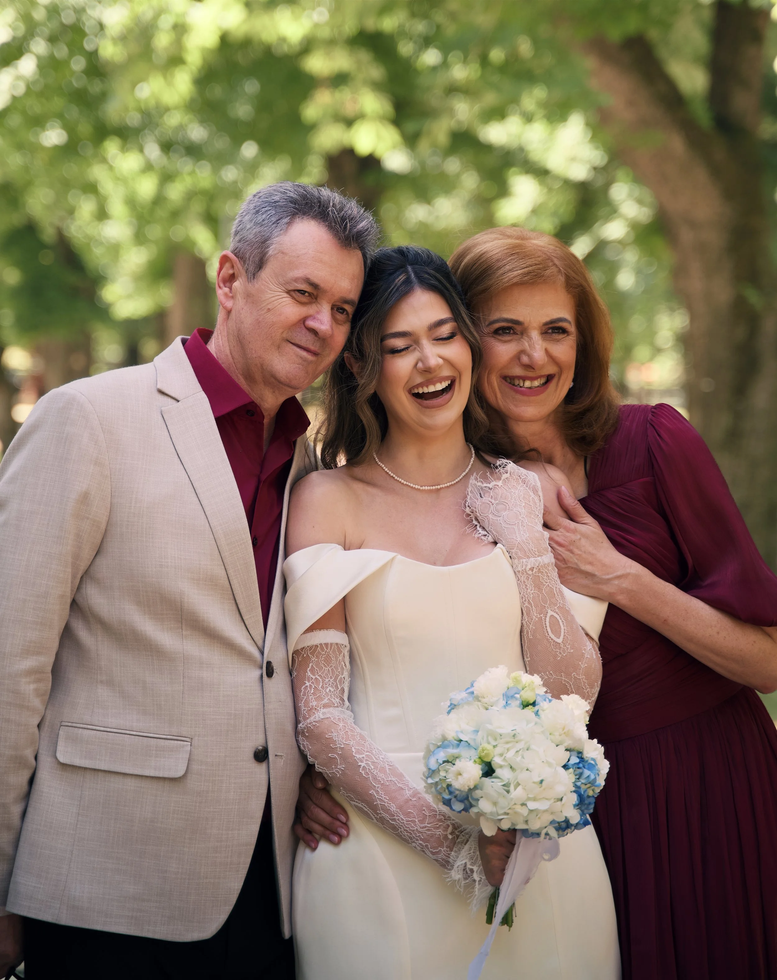 A woman in a wedding dress holding a bouquet, standing between a man and woman outdoors, all smiling and showing affection.