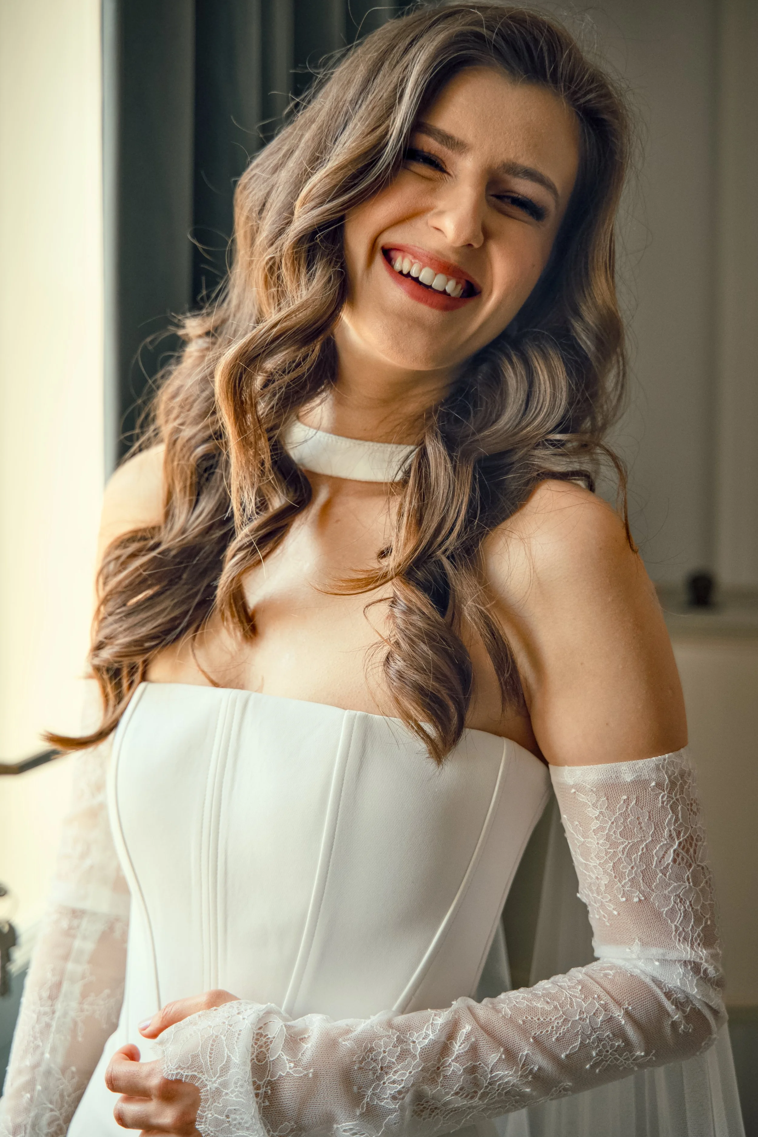 A young woman with long, wavy brown hair and makeup, wearing a white wedding dress with lace sleeves, smiling and winking.
