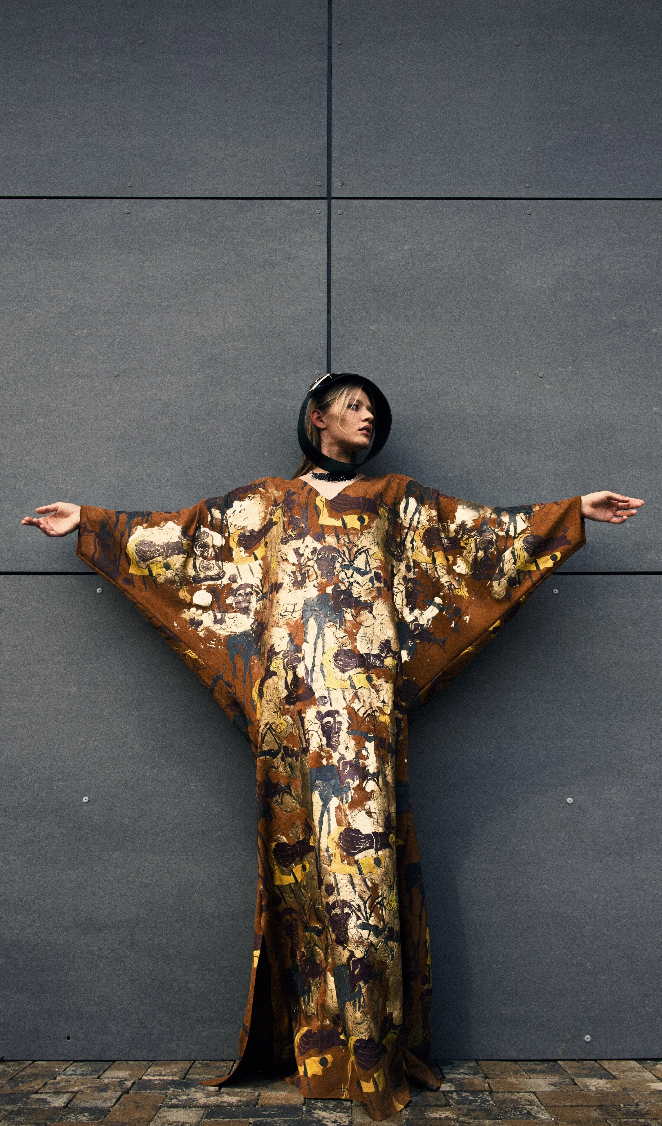 A woman standing against a gray wall with arms outstretched, wearing a long, colorful abstract dress and a black hat.
