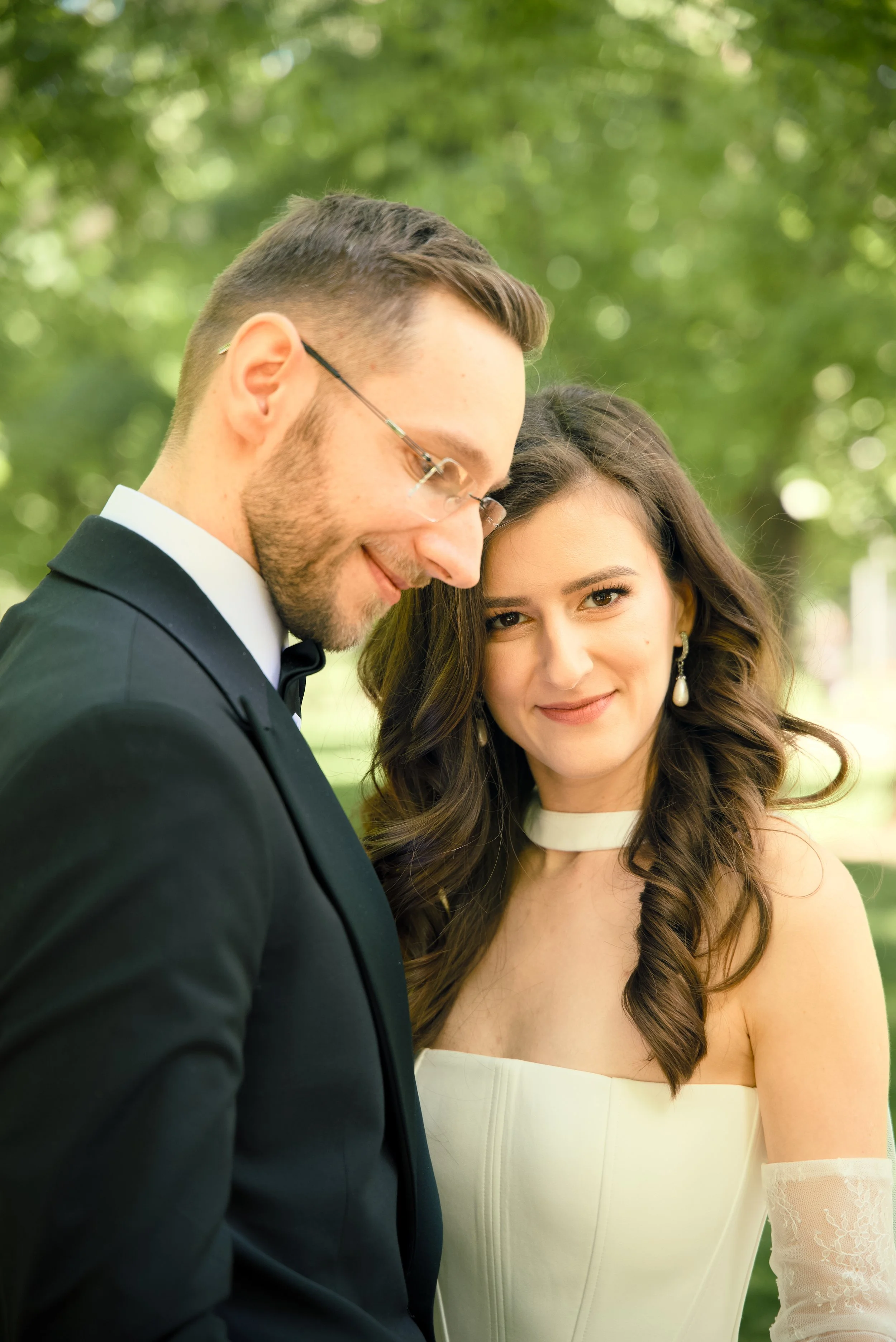 A couple dressed in formal wedding attire standing close together outdoors with blurred green trees in the background.