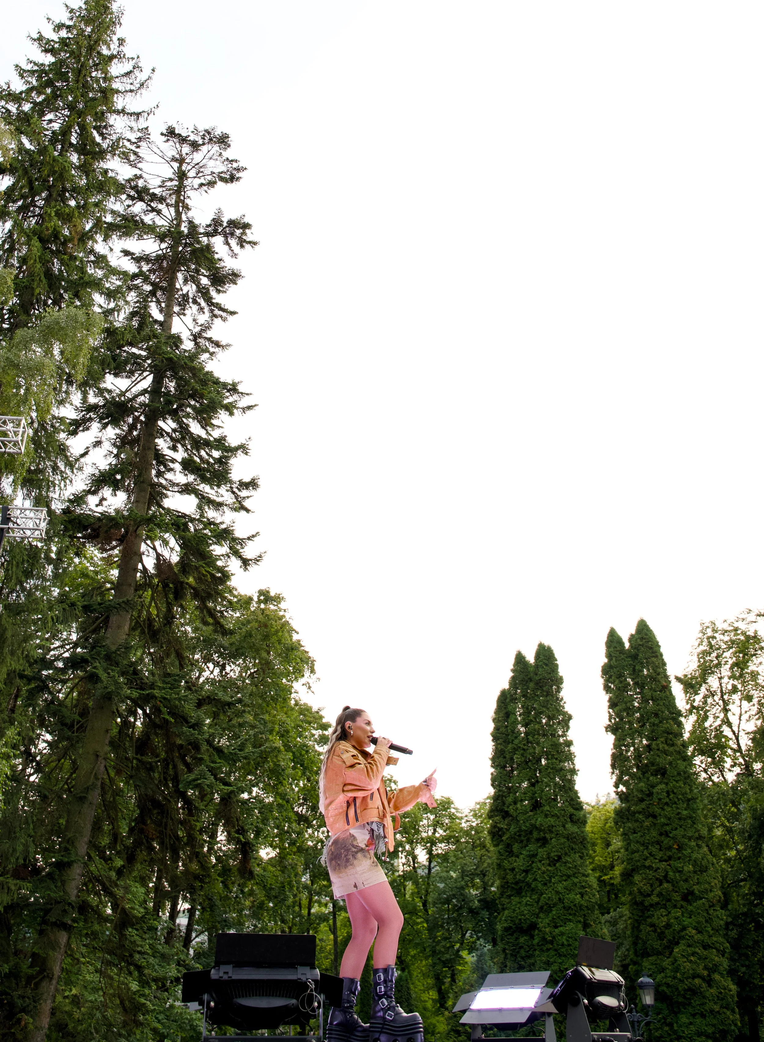 A female singer performing on an outdoor stage, holding a microphone in her right hand and a paper in her left hand. She is wearing a tan jacket, a skirt, pink tights, and black boots. There are tall trees and an overcast sky in the background.