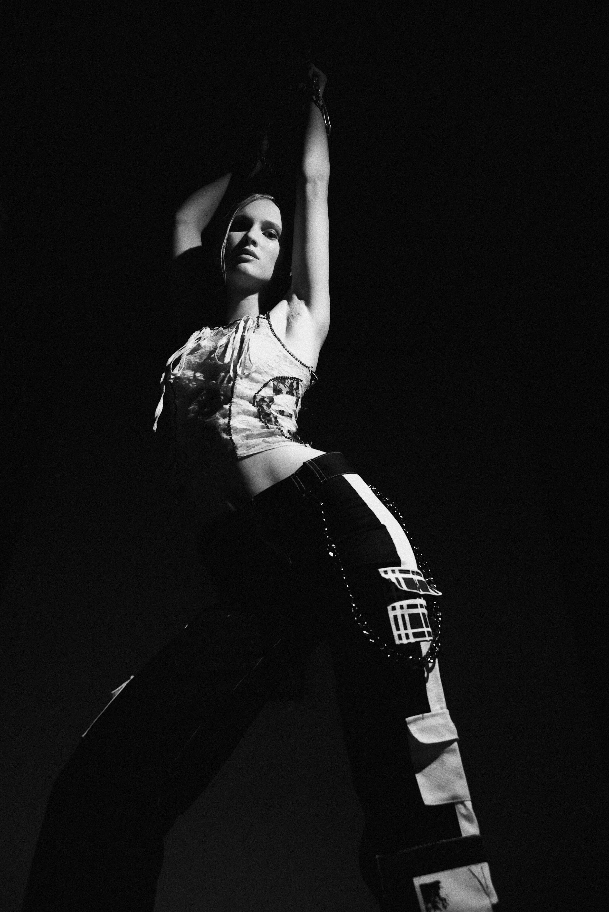 A woman with long hair posing with her arms raised, wearing a patterned sleeveless top and pants with patches, in a high-contrast black and white photograph.