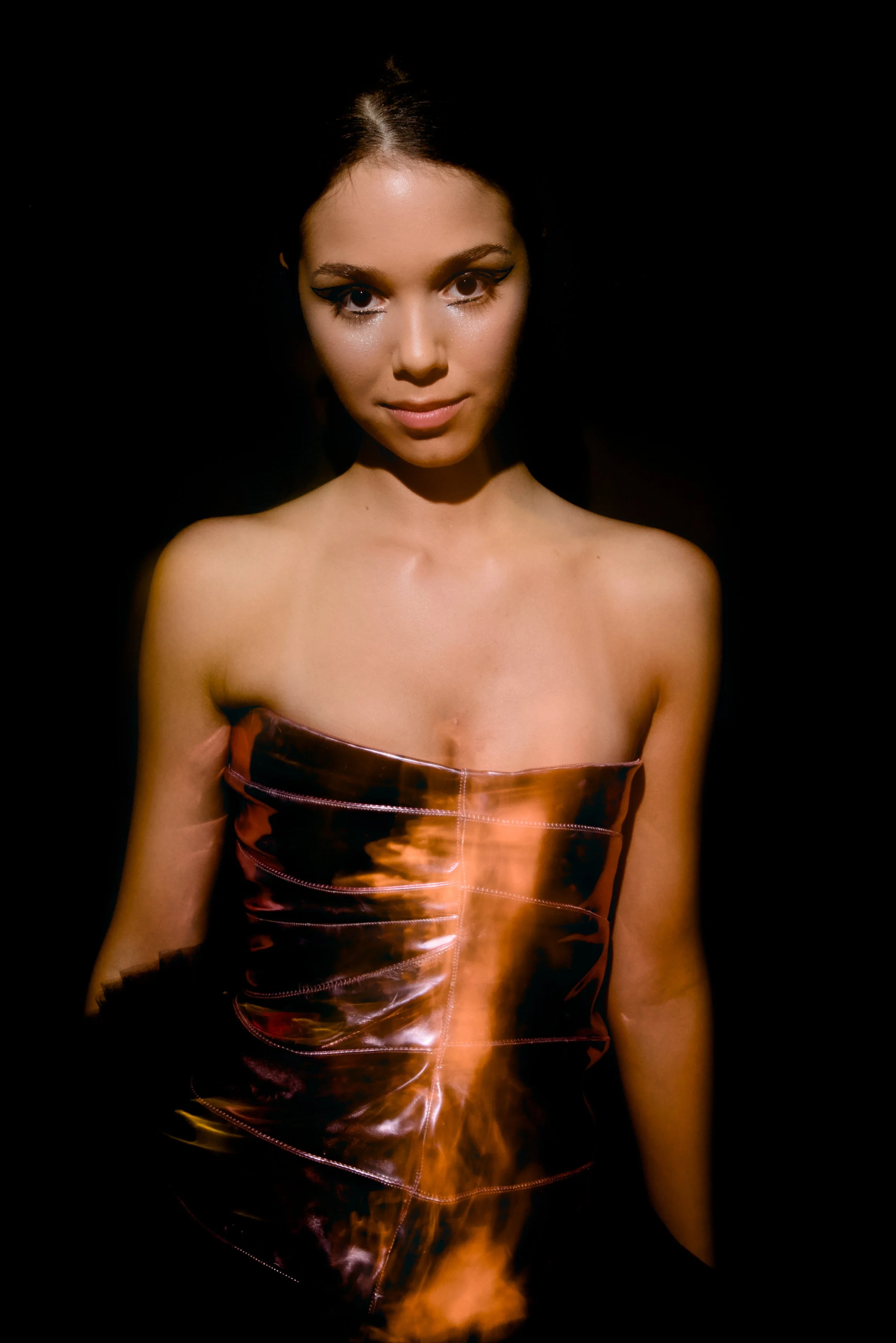 A woman with dark hair and makeup, wearing a strapless, shiny, burnt orange-colored dress against a black background.