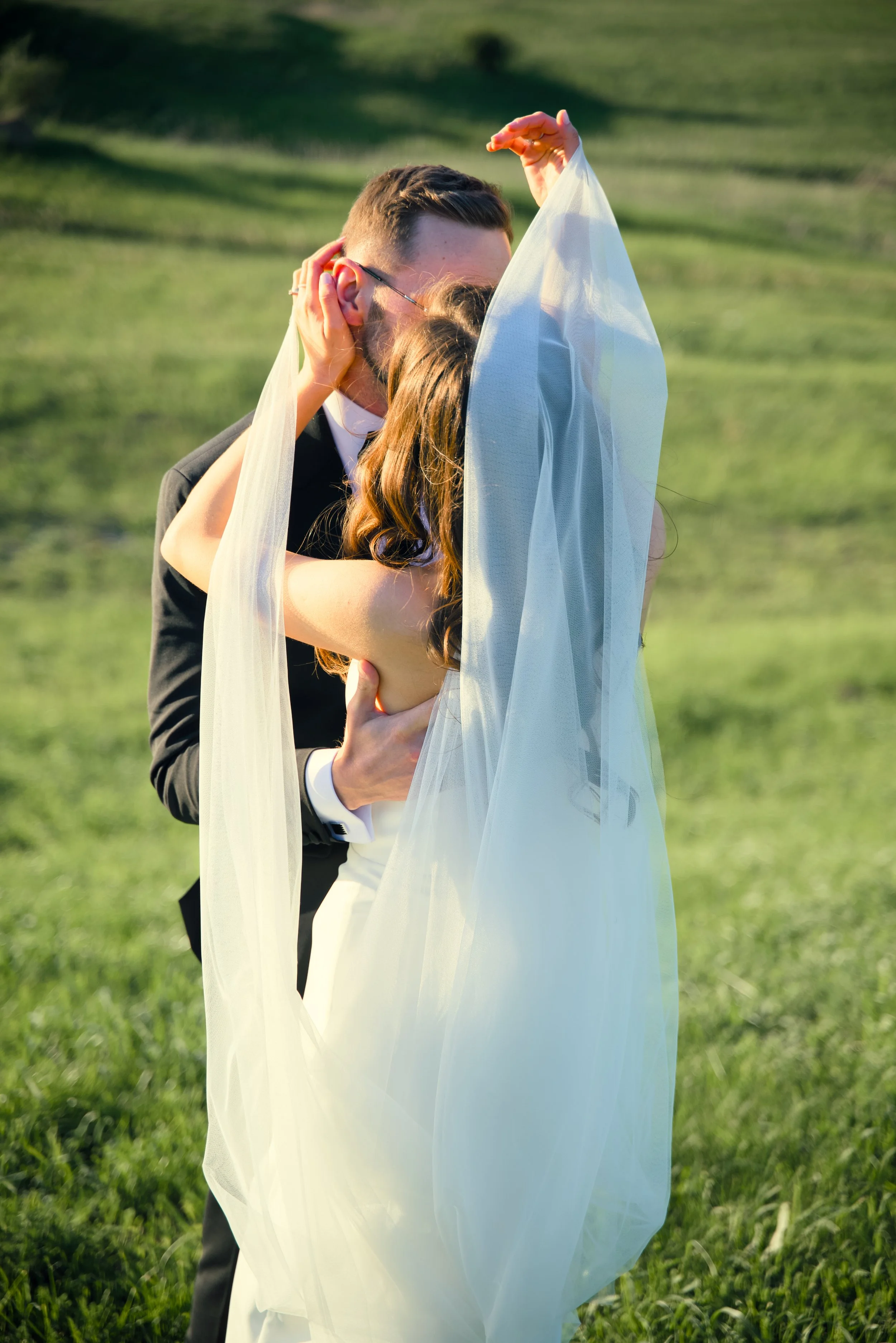 A bride and groom share a kiss outdoors with a veil flowing over them.