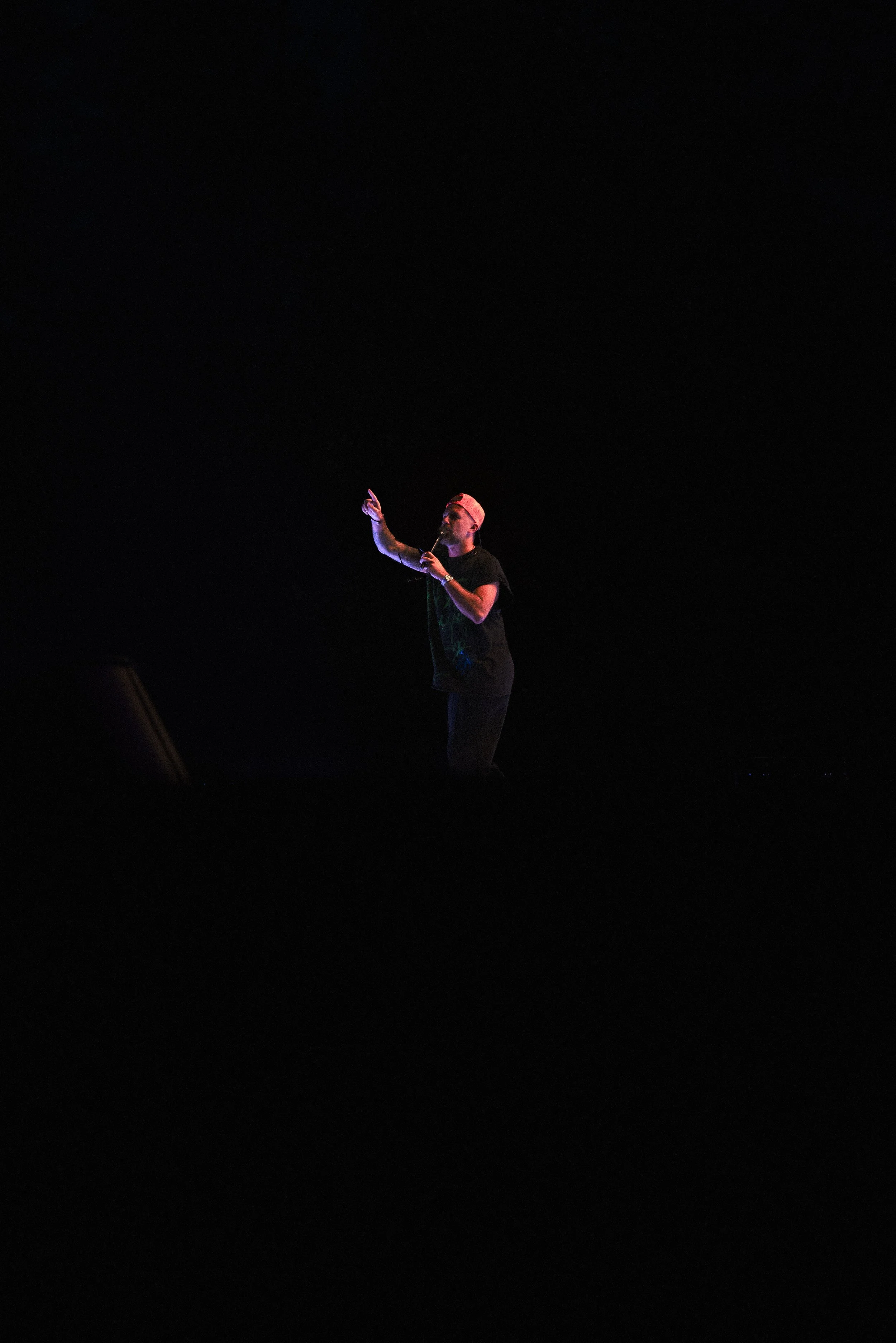 A person wearing a black shirt and a backward cap, holding a microphone, standing on a dark stage with minimal lighting.