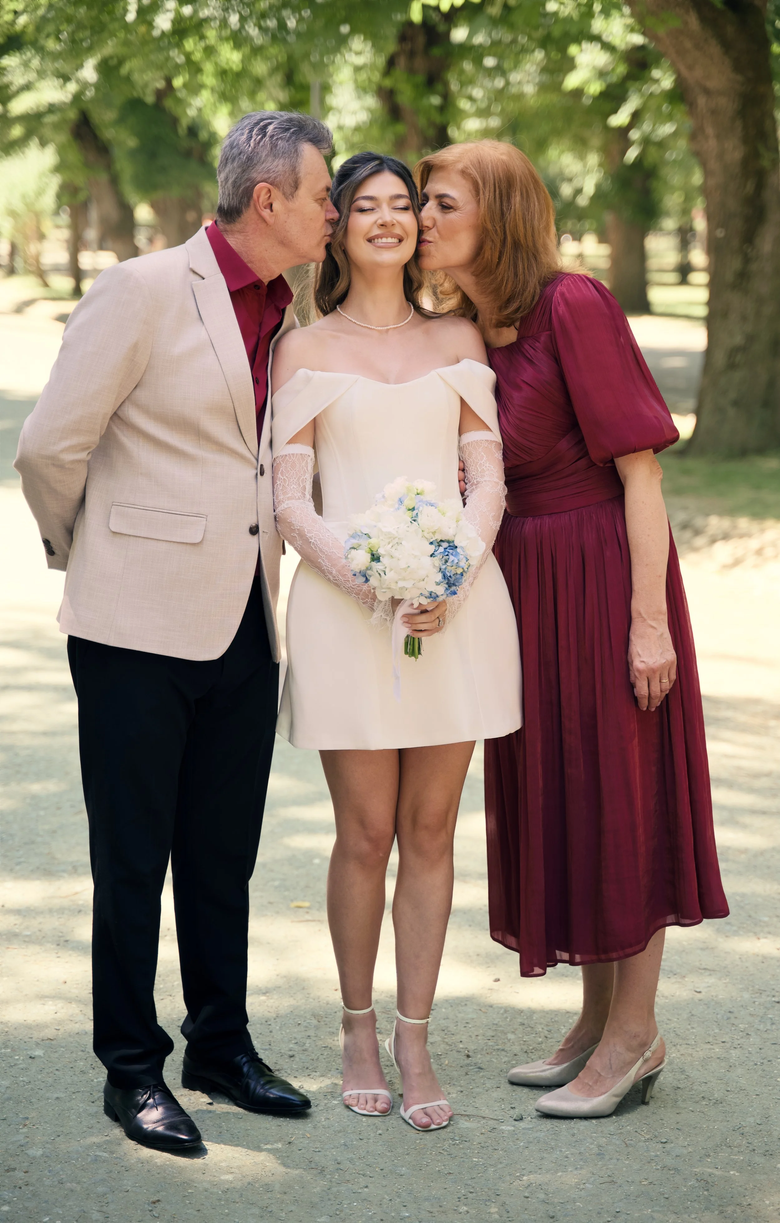 Young woman in a white dress holding a bouquet, standing between a man and a woman, outdoors with trees, both adults kissing her on the cheeks