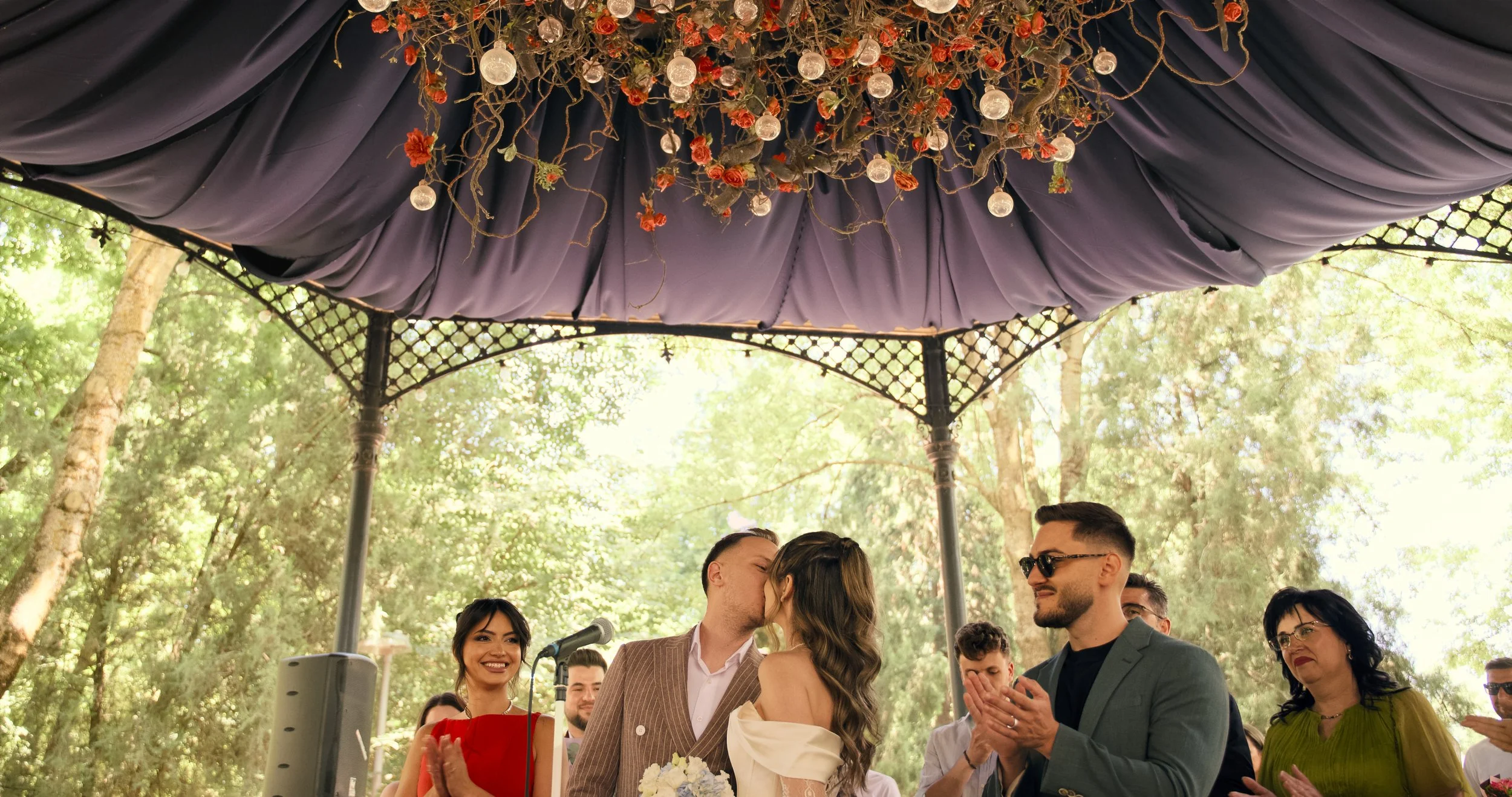 Couple shares a kiss during their wedding ceremony under a decorated outdoor canopy with guests clapping and smiling.