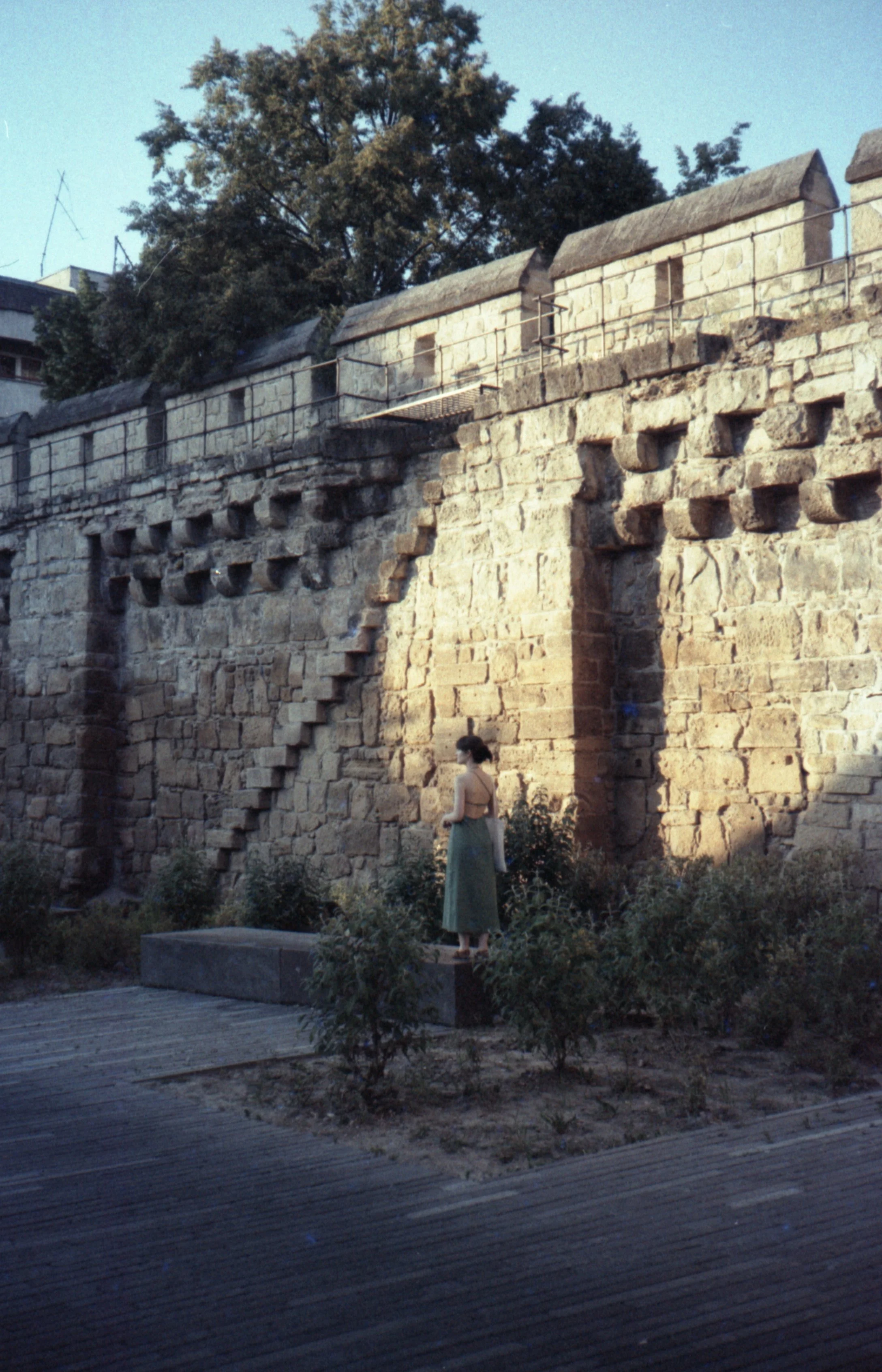 A woman standing on a stone block in front of an old stone fortress wall with stairs and a railing.