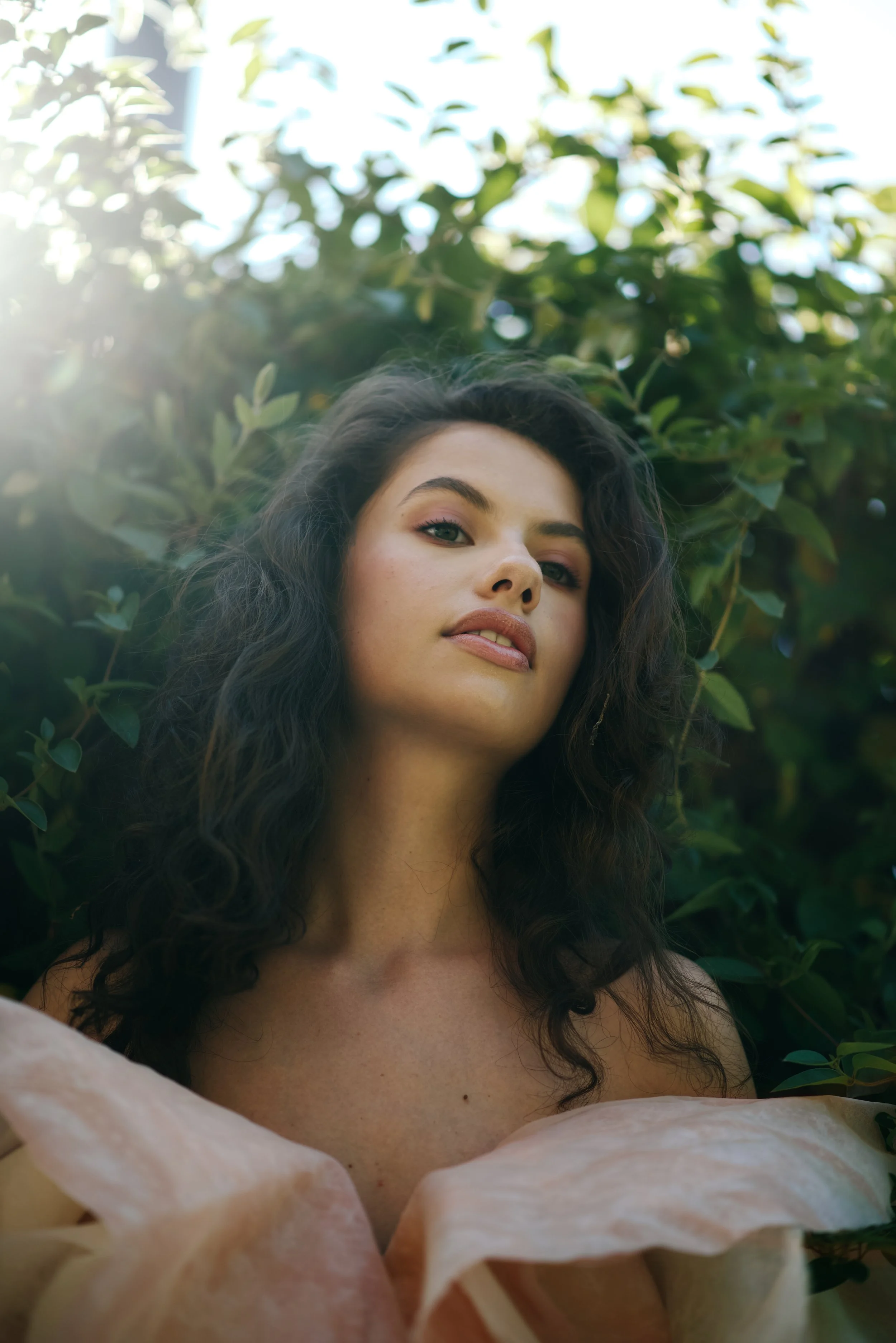 A woman with wavy dark hair is outdoors, standing in front of green foliage, looking into the camera with a confident expression, sunlight filtering through the trees.