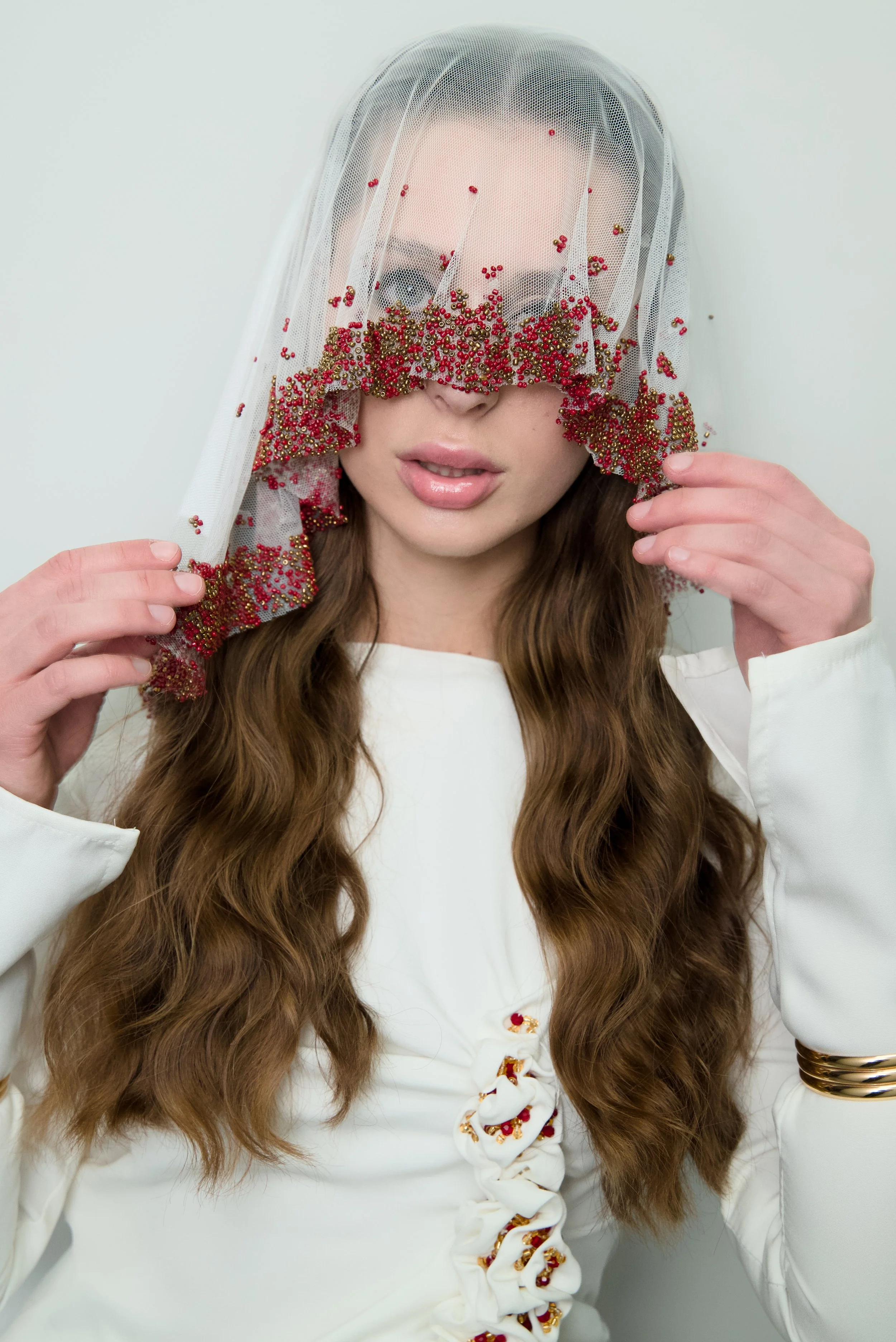 A woman with long wavy brown hair wearing a white dress with a ruched detail and gold jewelry. She has a sheer veil or netting with red and gold embellishments draped over her face, partially covering her eyes and mouth.