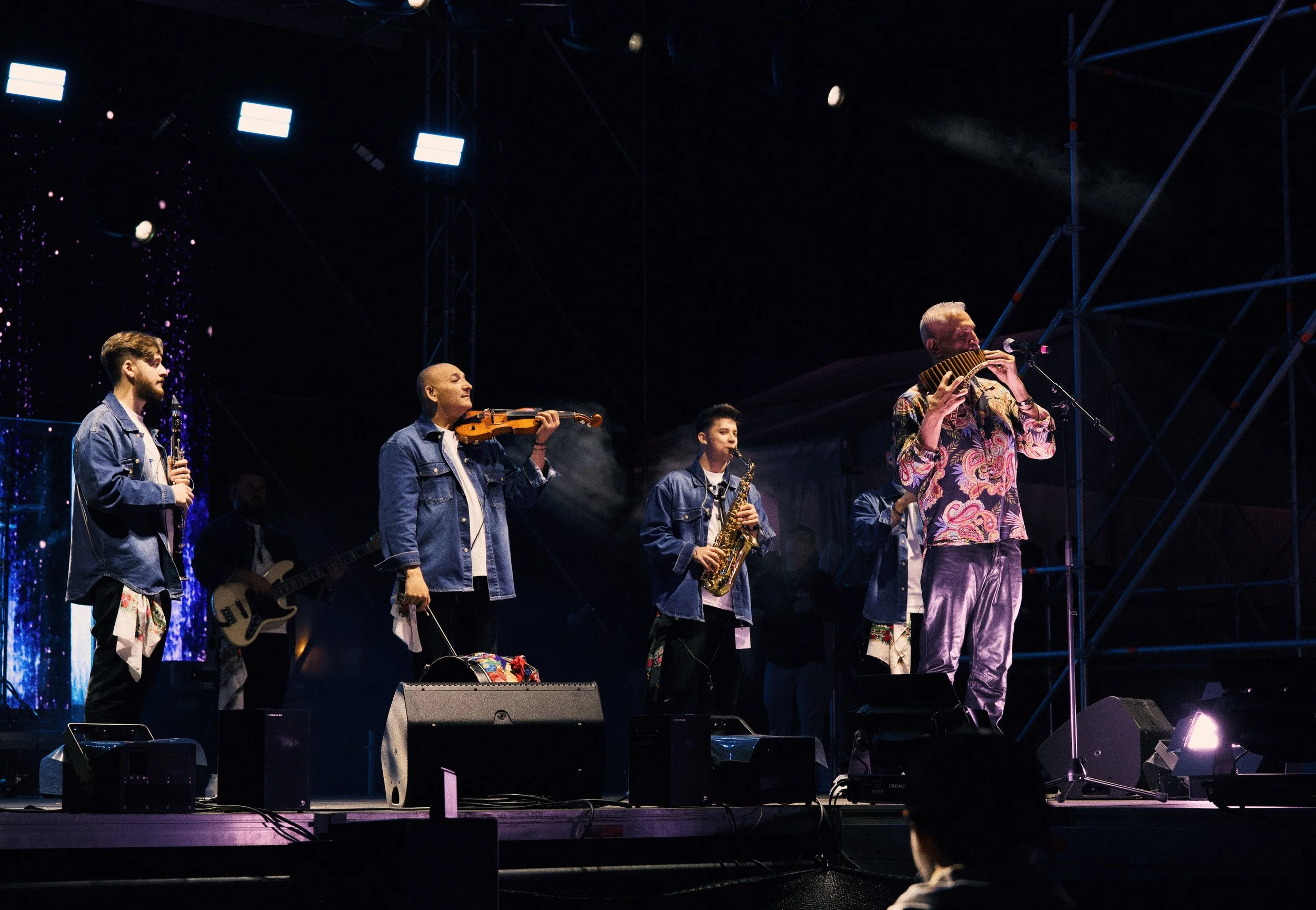 A group of musicians performing on stage with various instruments, including violins, saxophone, wind instrument, and bass guitar, under stage lighting.