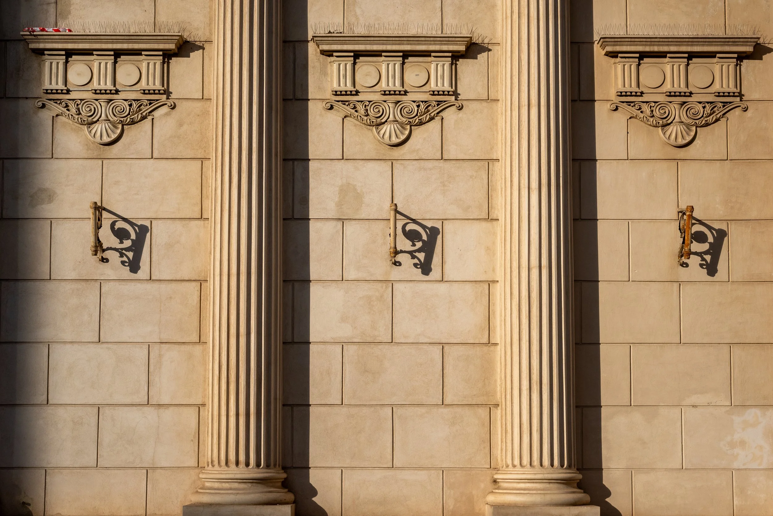 Close-up of a beige stone building facade with decorative classical columns, ornate corbels, and two wall-mounted vintage-style lamps casting shadows.