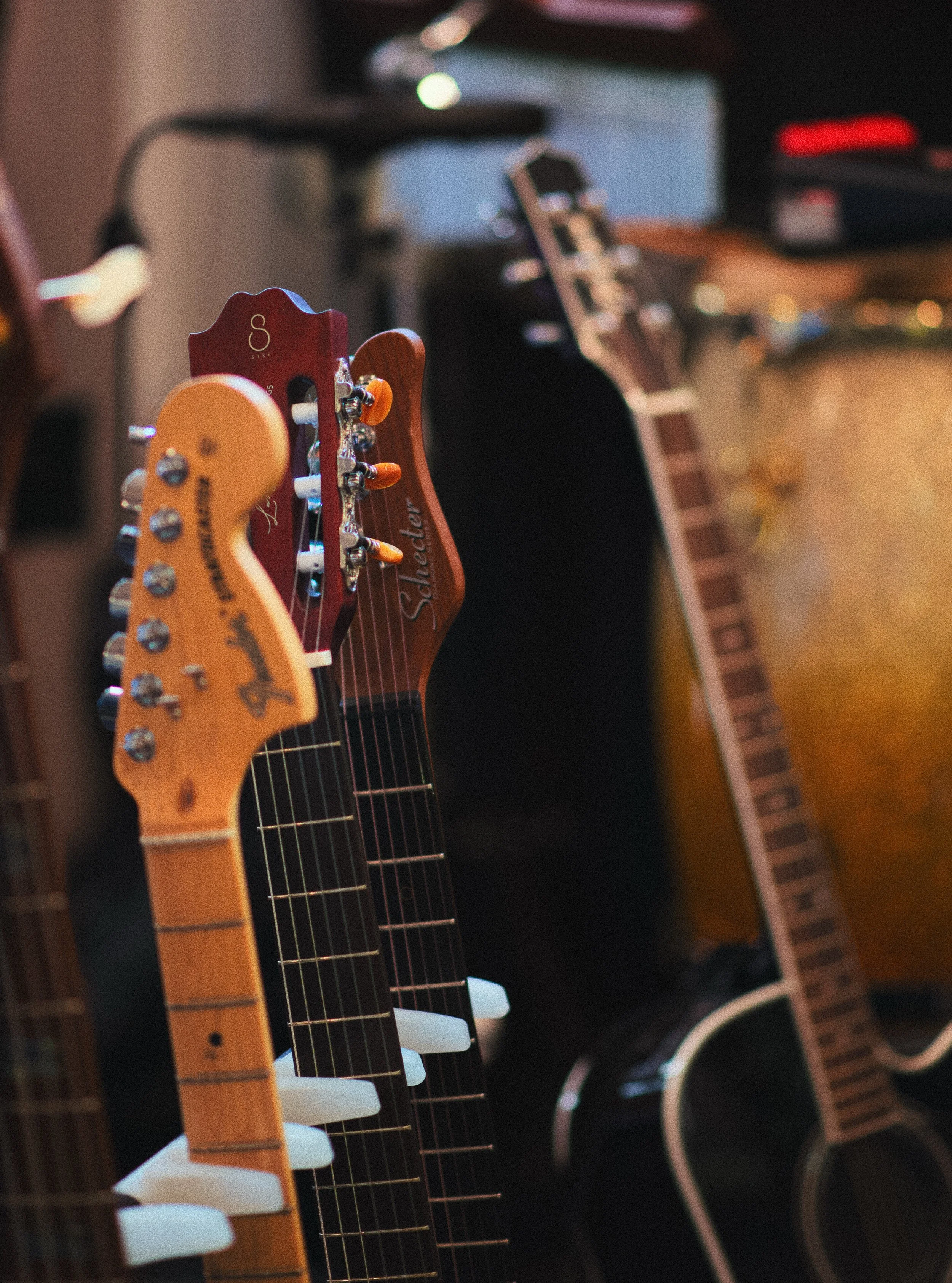 Close-up of multiple guitars with headstocks and necks, including Fender and Schecter brands, against a blurred background of musical equipment.