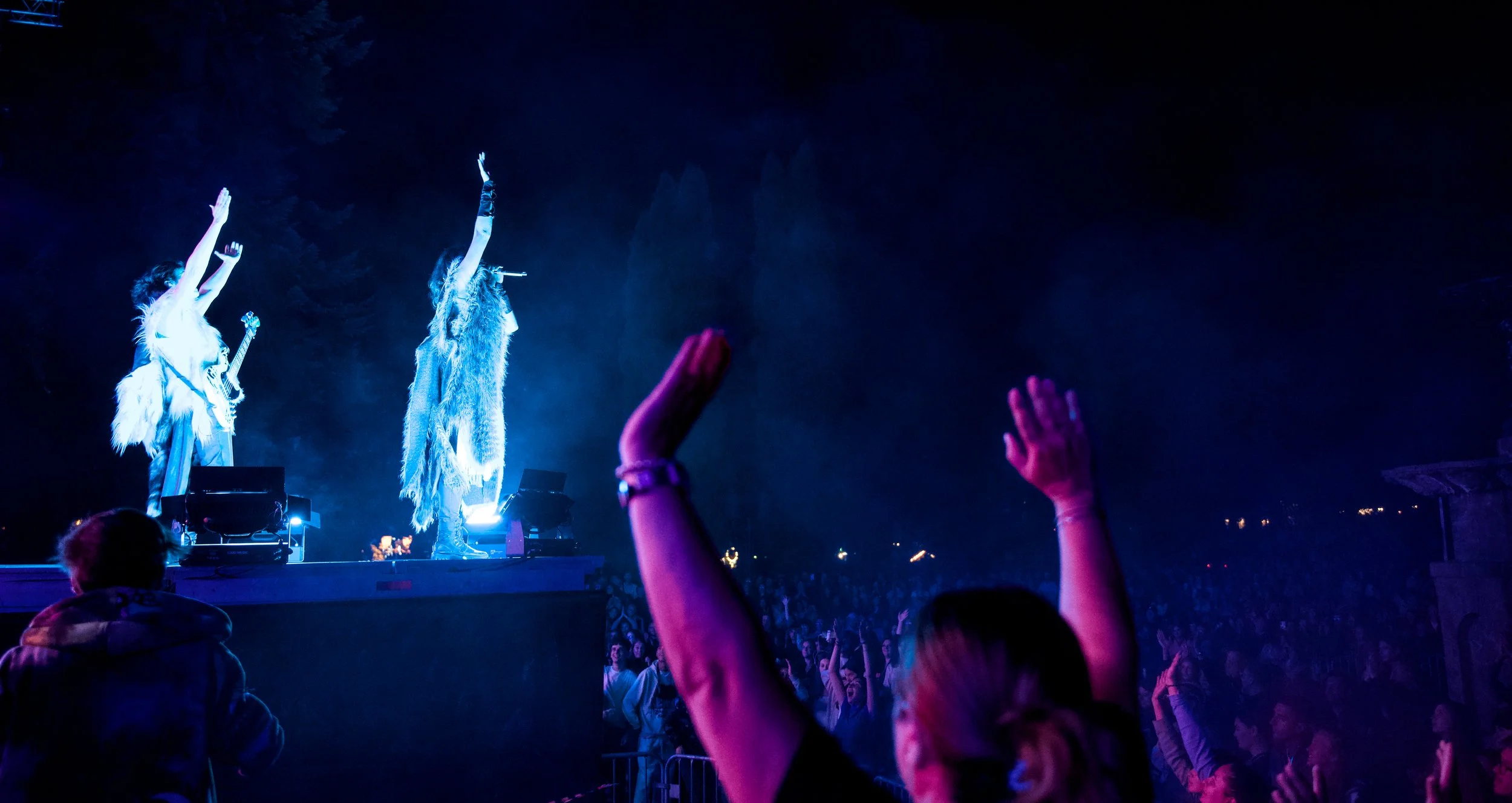 Two performers on stage at night with arms raised, audience with hands up, blue and purple stage lighting, outdoor setting.