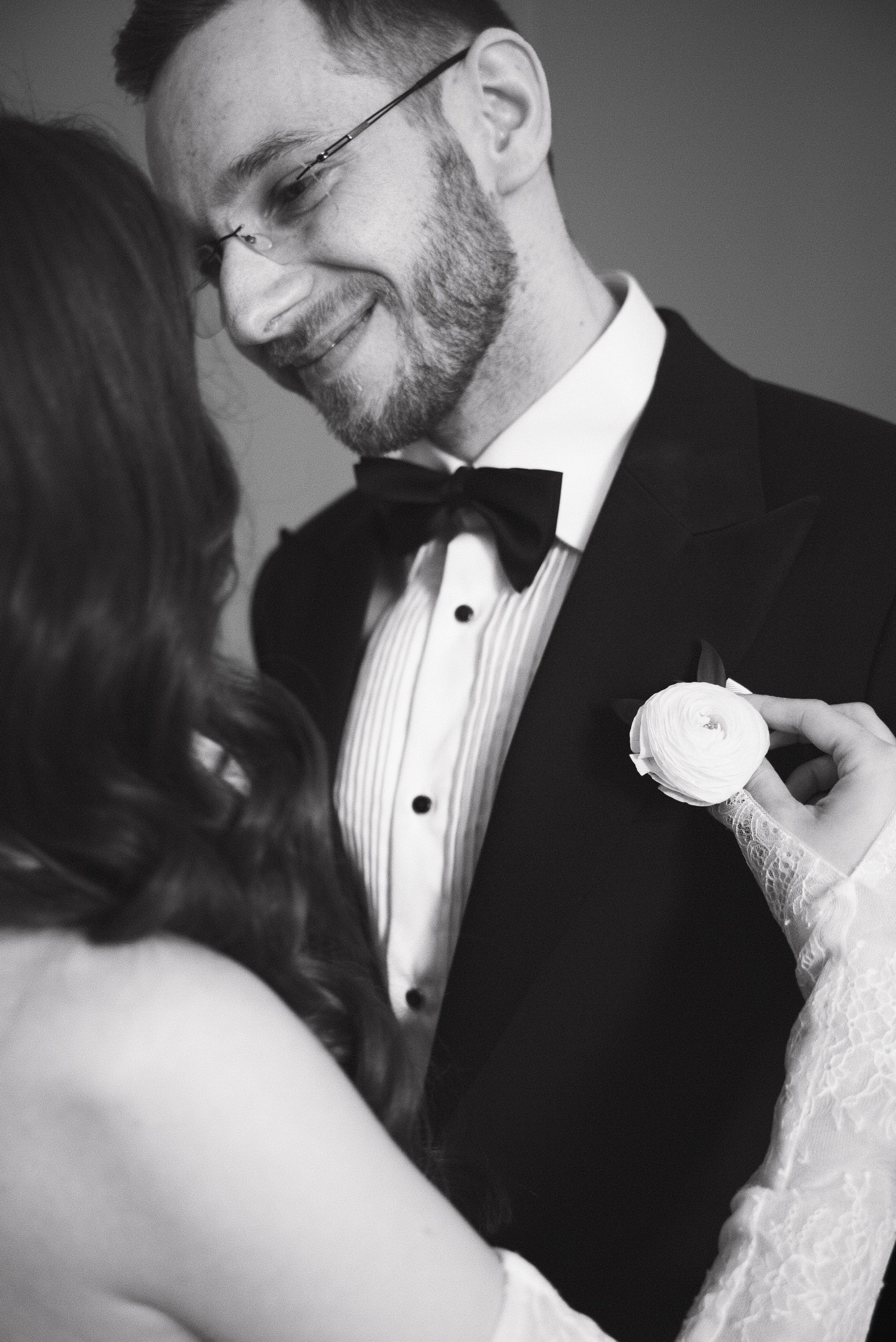 A couple dressed in formal wedding attire, with the woman pinning a flower on the man's tuxedo.