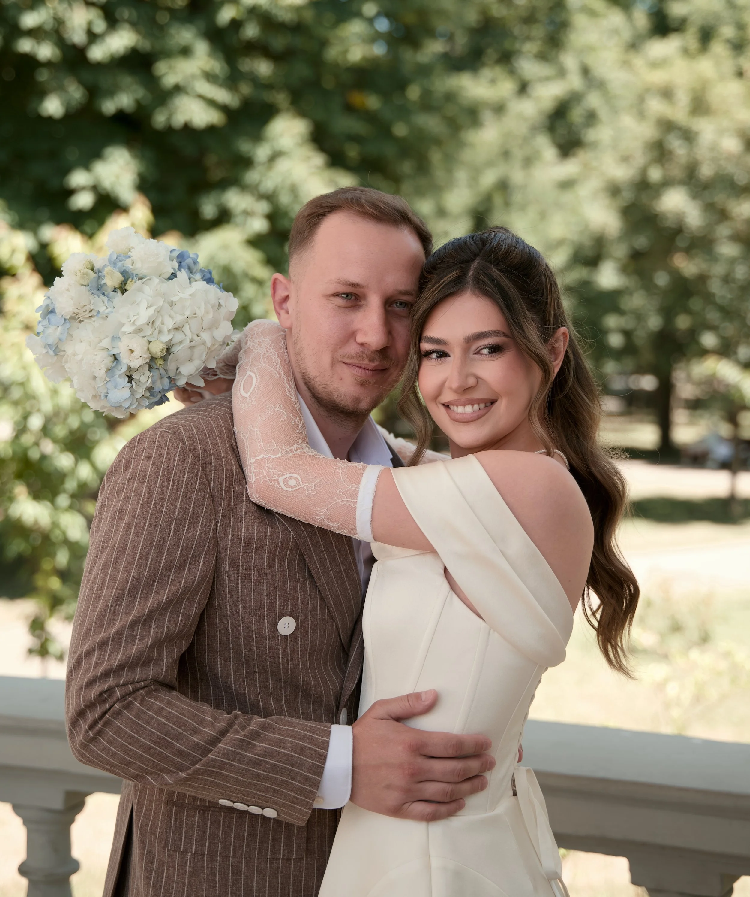 A happy couple in wedding attire, with the woman holding a bouquet of white and light blue flowers, embracing outdoors against a backdrop of green trees and sunlight.