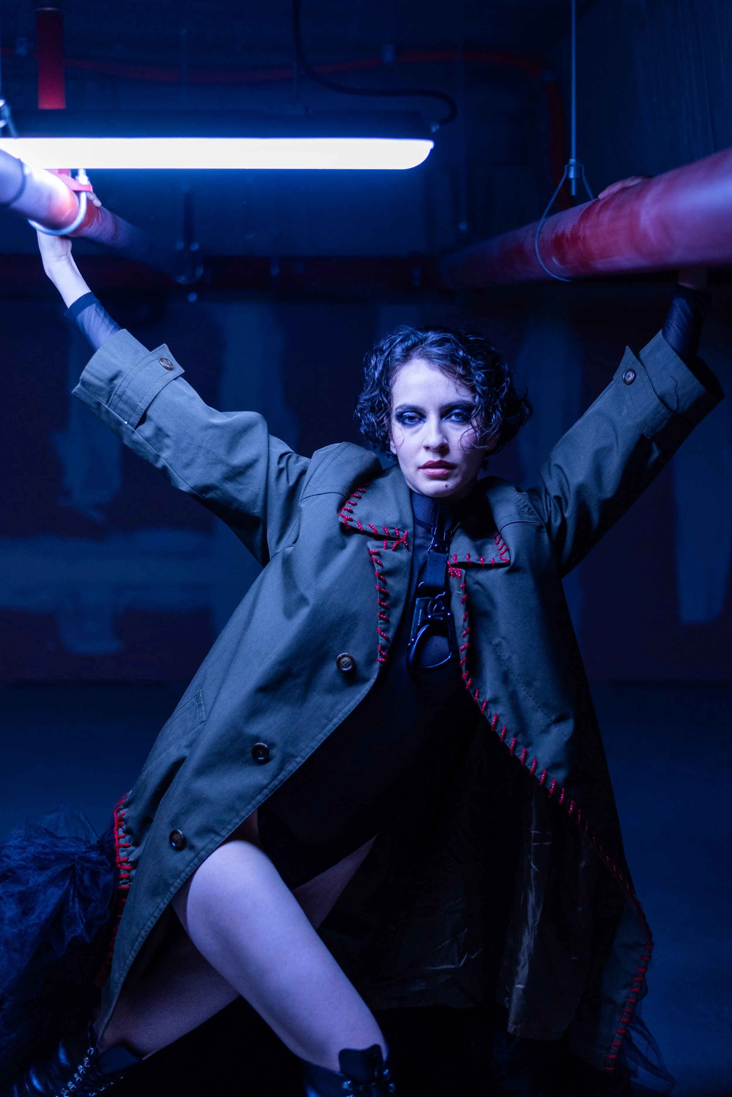 A woman with short curly hair poses under blue/white lighting in a dark industrial setting. She wears a green coat with red stitching, black lace-up boots, and has a serious expression.