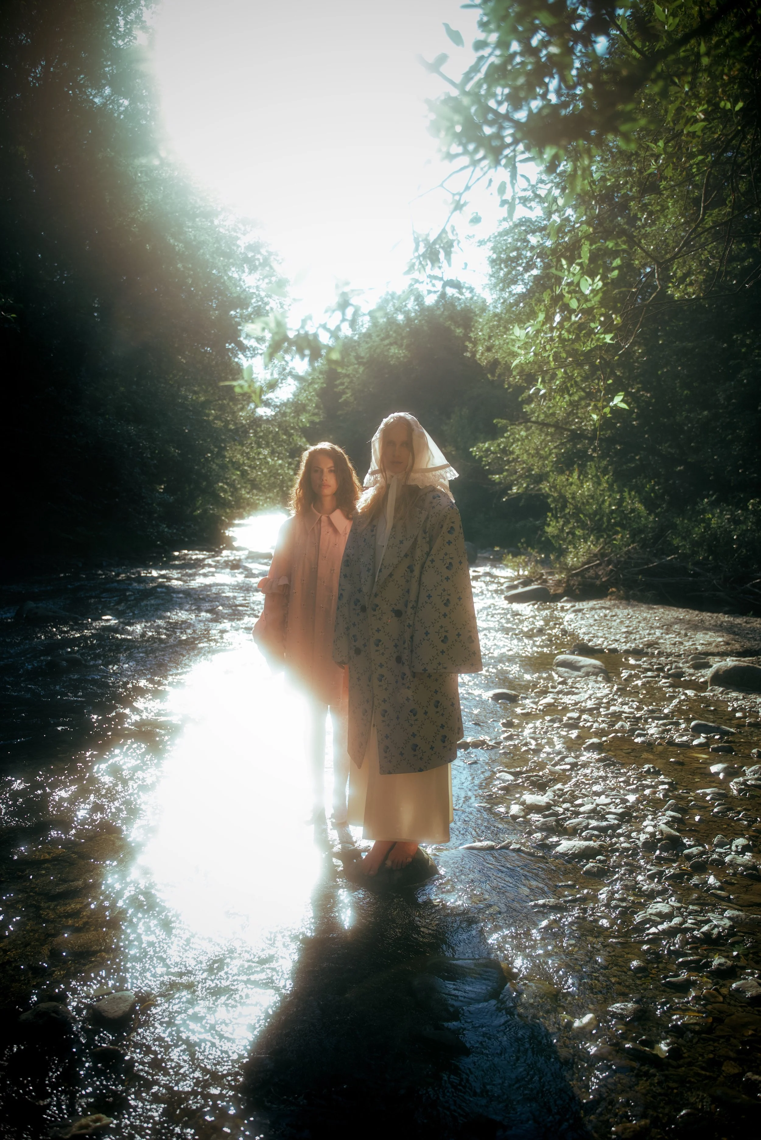 Two women in vintage dresses standing in a shallow, rocky creek with sunlight filtering through trees overhead.