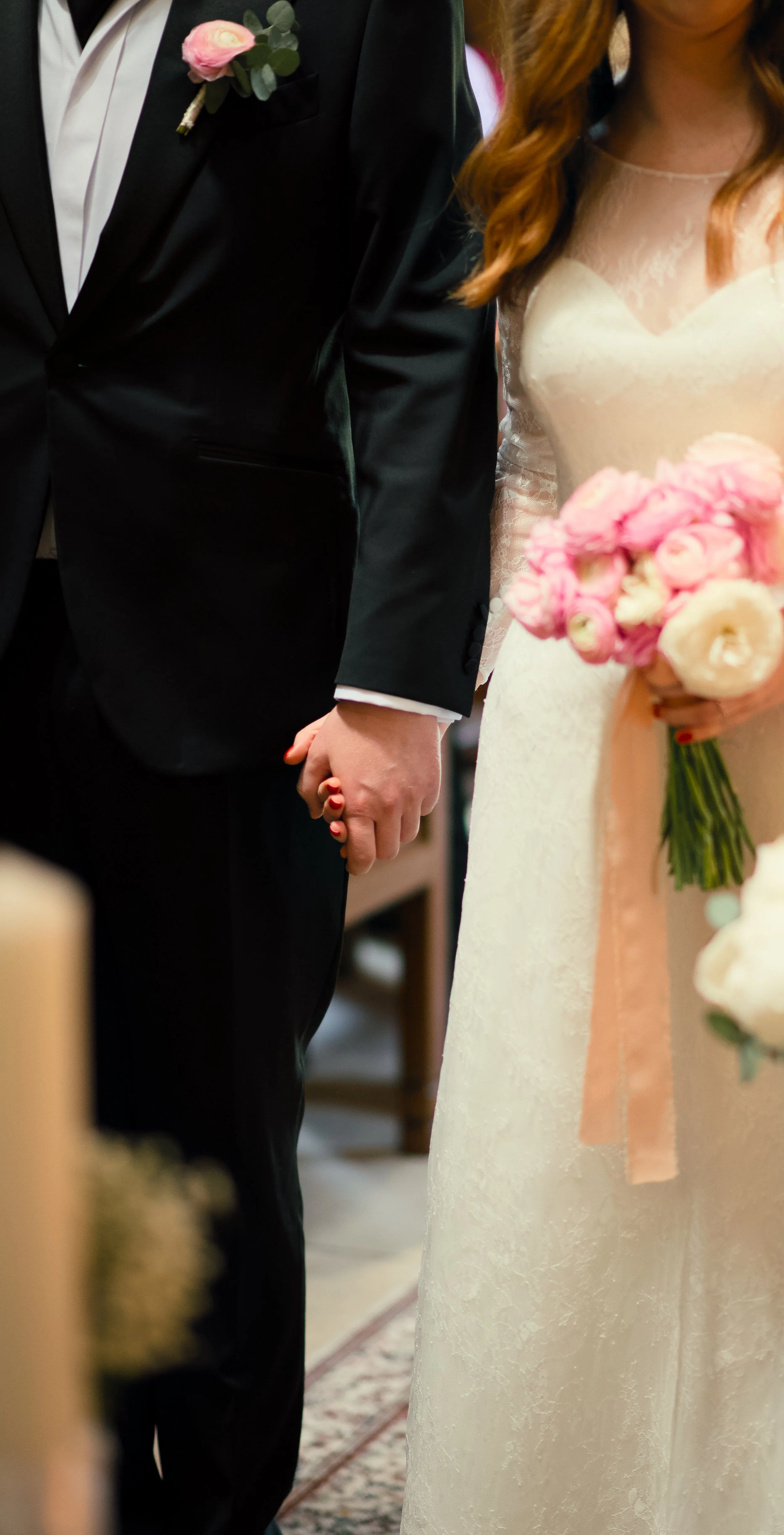 A bride and groom holding hands during a wedding ceremony. The bride is dressed in a white lace wedding gown and holding a bouquet of pink and white flowers. The groom is wearing a black tuxedo with a white shirt and a pink boutonniere.