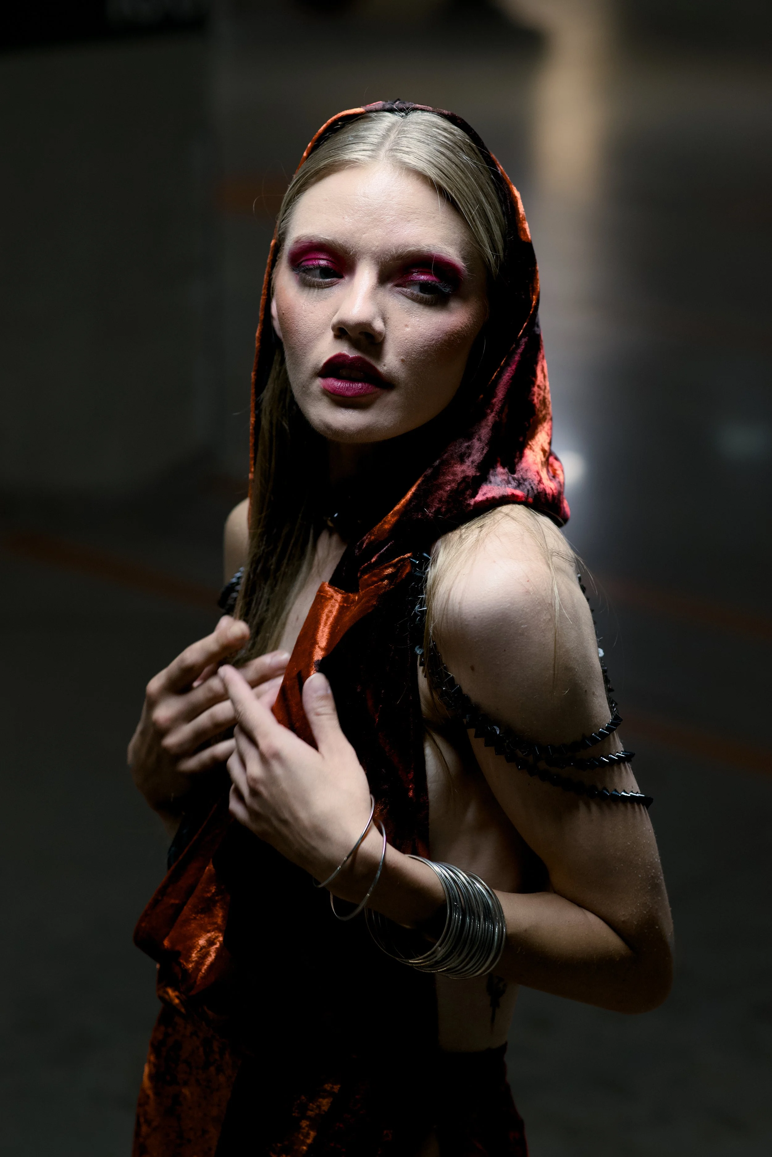 A woman with long blonde hair wearing a burnt orange velvet hooded top, dark makeup with pink eyeshadow, and multiple silver bangles on her wrists, posing with a moody expression in a dark, blurred background.