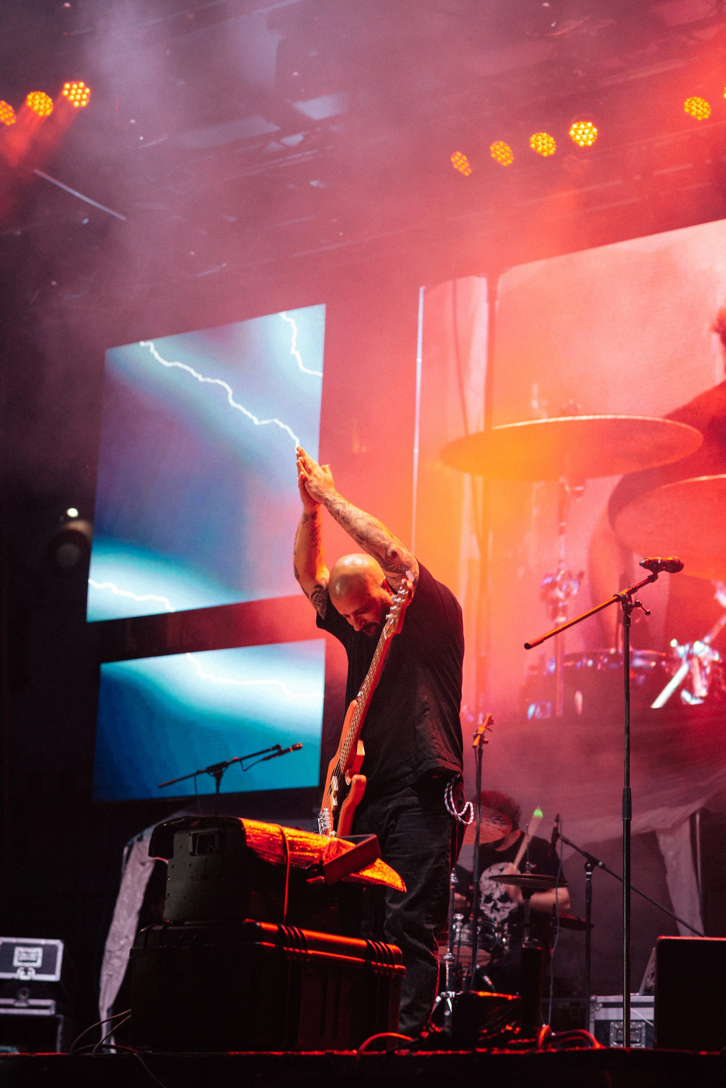A bald musician with tattoos wearing black clothing on stage, playing a guitar with hands raised together, illuminated by red and orange stage lights, with electronic equipment and a large screen displaying lightning in the background.