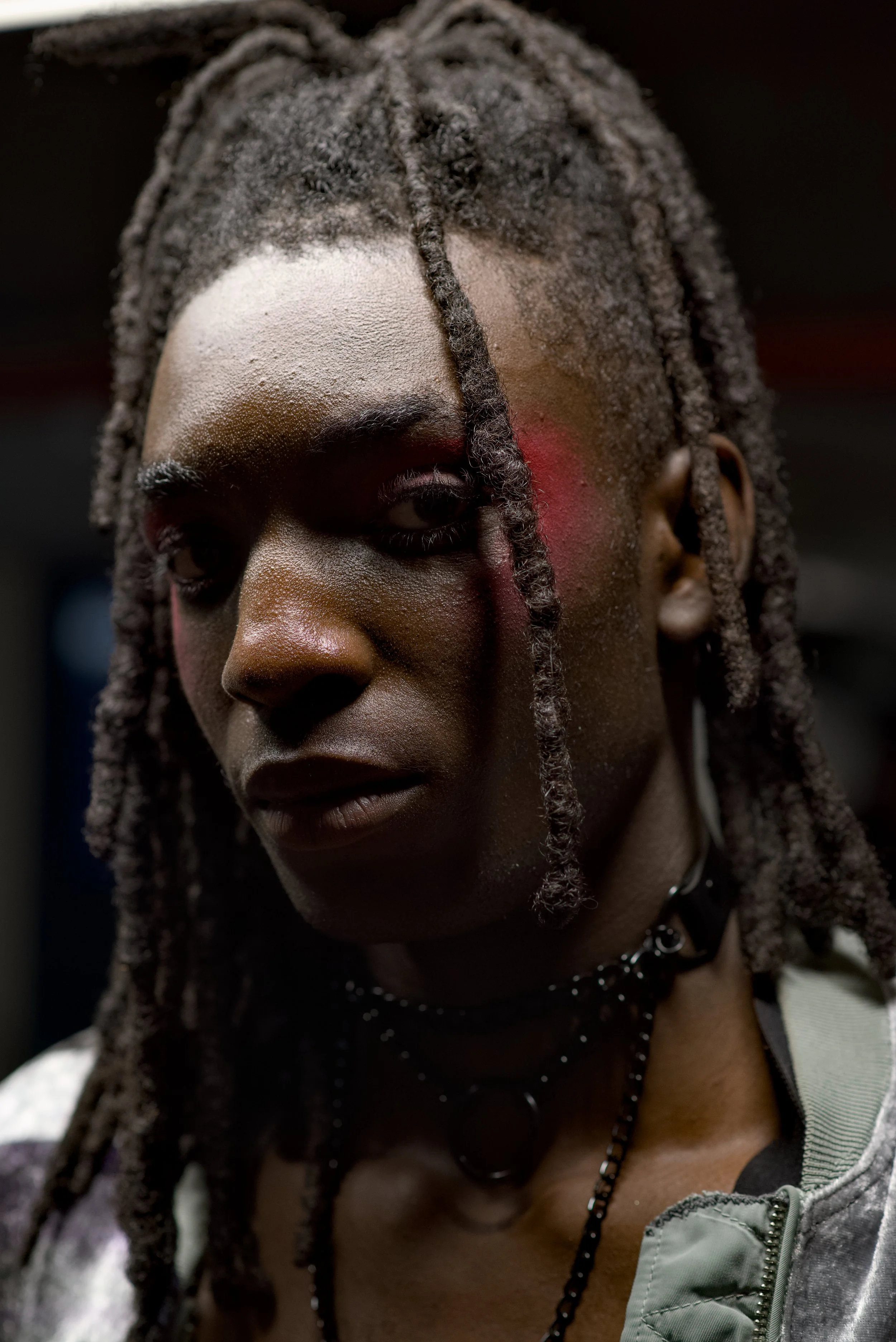 Close-up of a person with dreadlocks, dark skin, and makeup, wearing layered necklaces and a grey jacket.