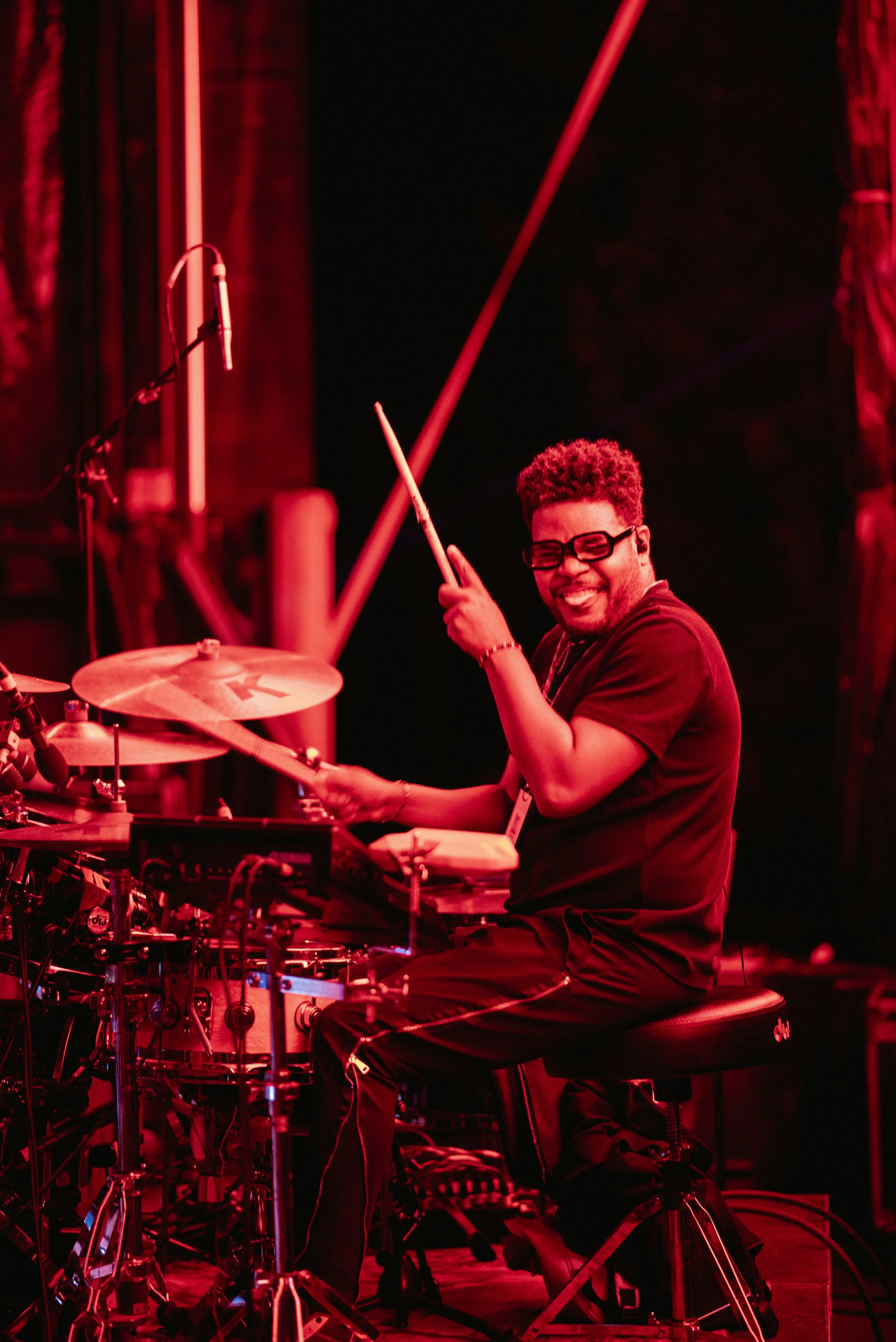A smiling drummer playing a drum set on stage under red lighting.
