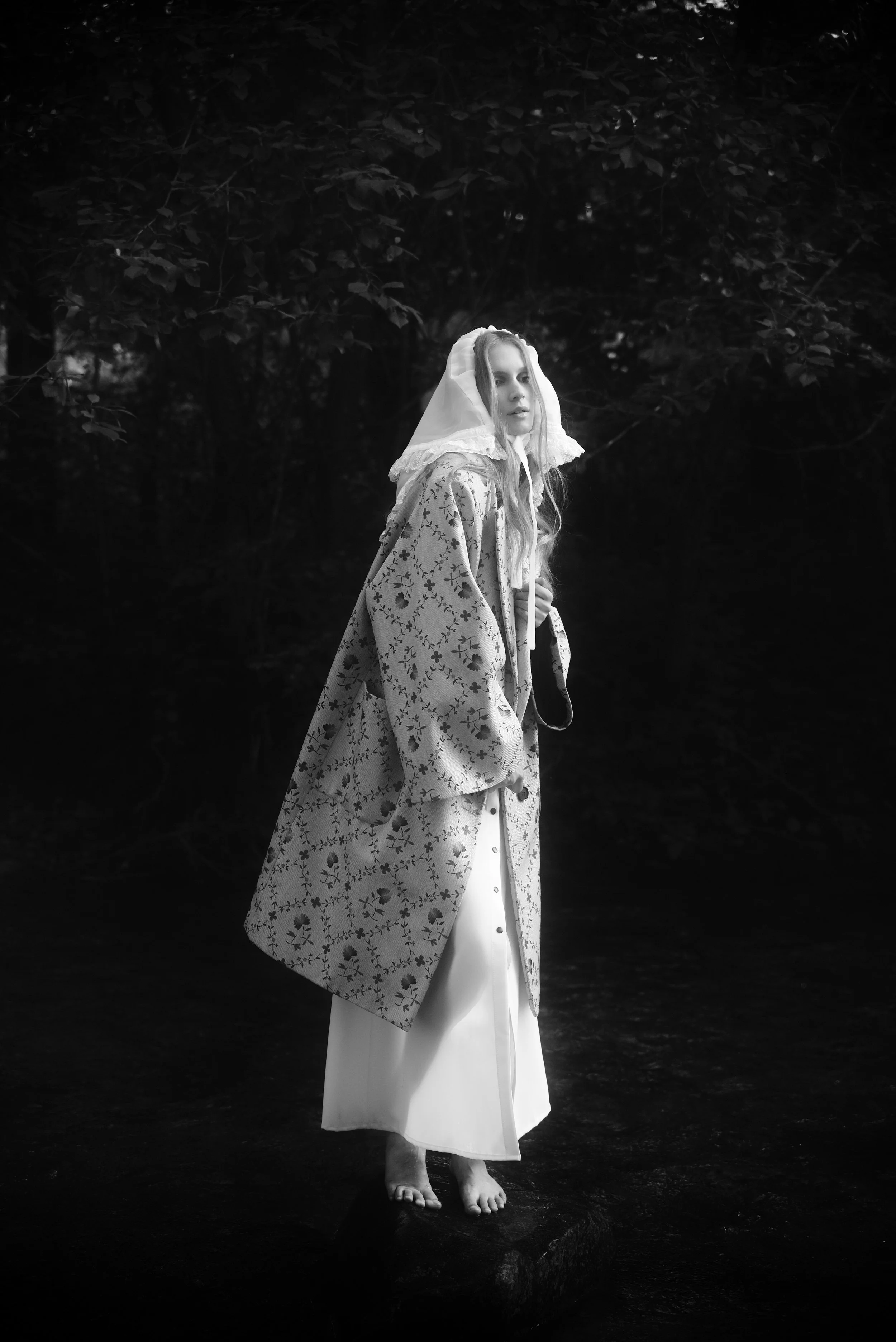 A woman dressed in a vintage-style outfit, standing barefoot on a rock near water, with dark foliage in the background, captured in black and white.