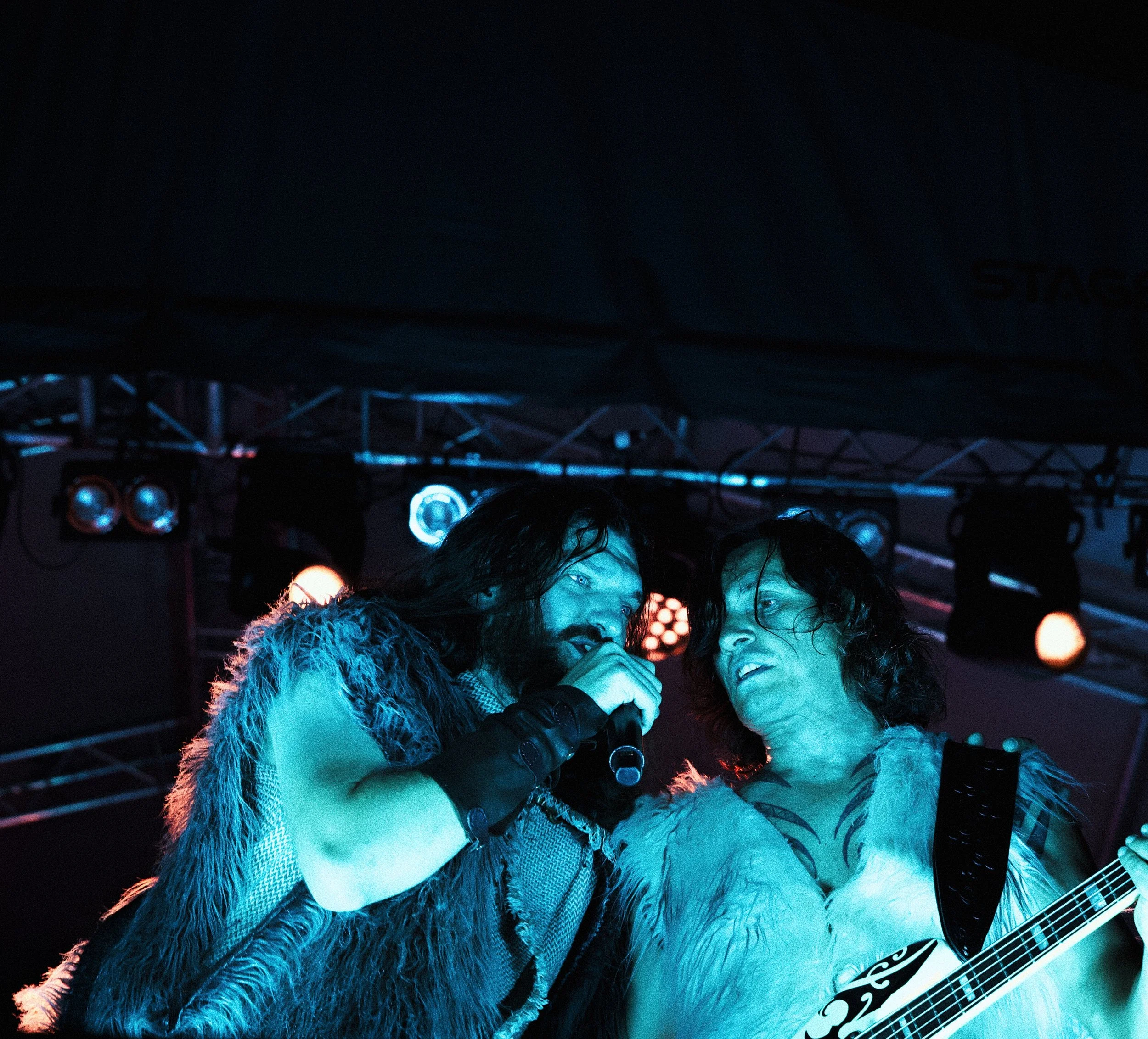 Two musicians performing on stage, one singing into a microphone and the other playing a guitar, under stage lights with a dark background.