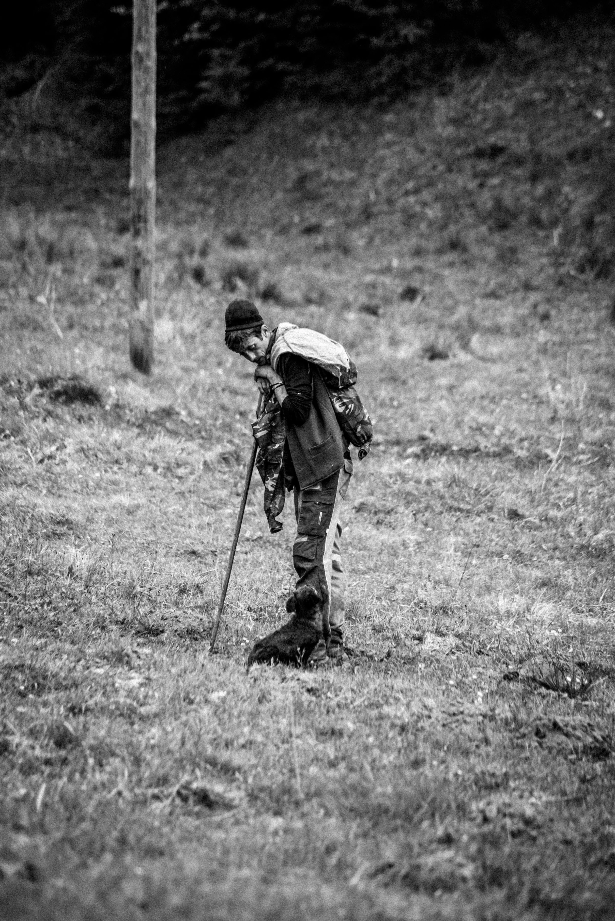 A person with a backpack and coat standing on grassy terrain with a small dog sitting at their feet, outdoors on a cloudy day.
