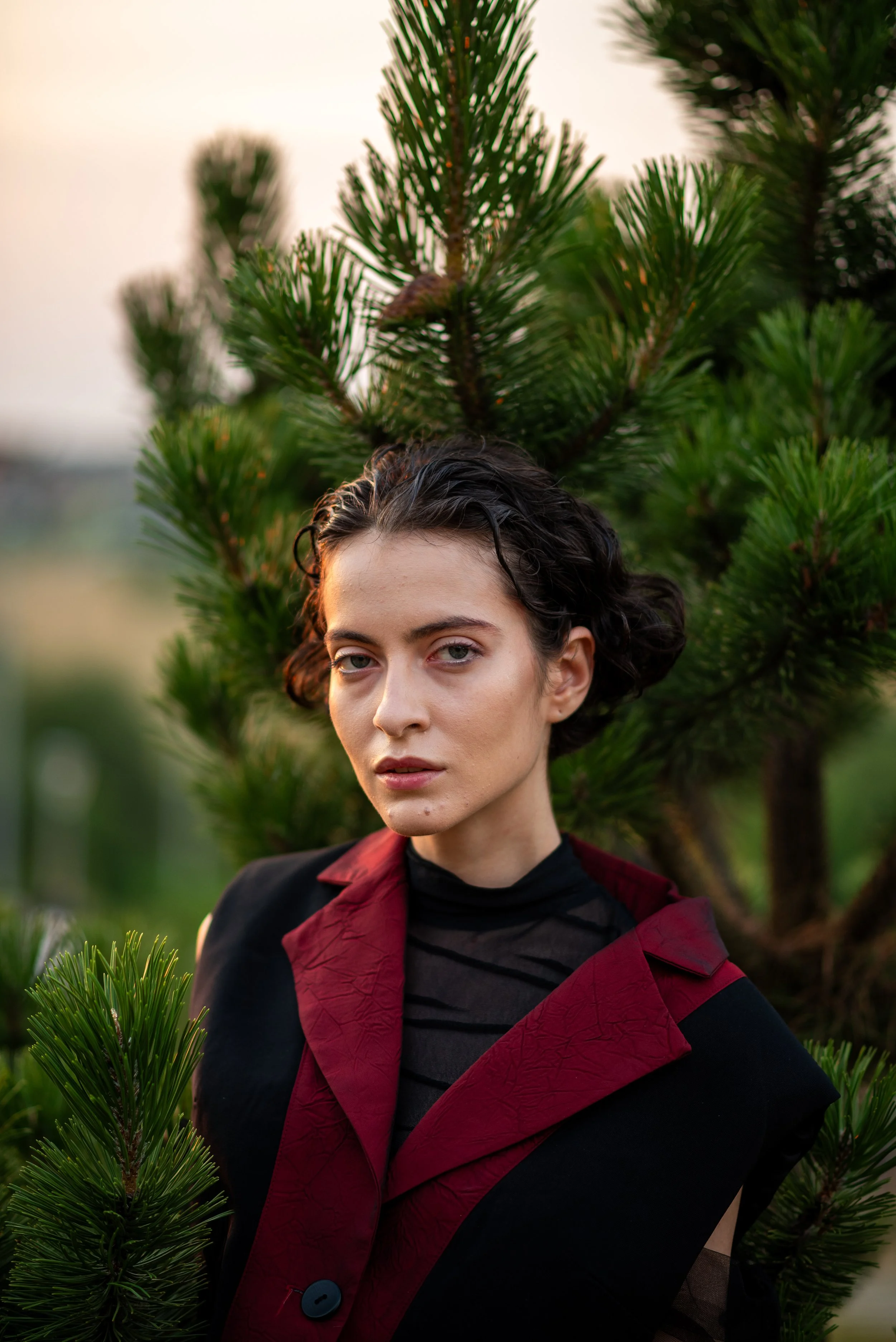 A woman with short dark hair and piercing gaze stands outdoors in front of a green pine tree, wearing a black and red jacket.
