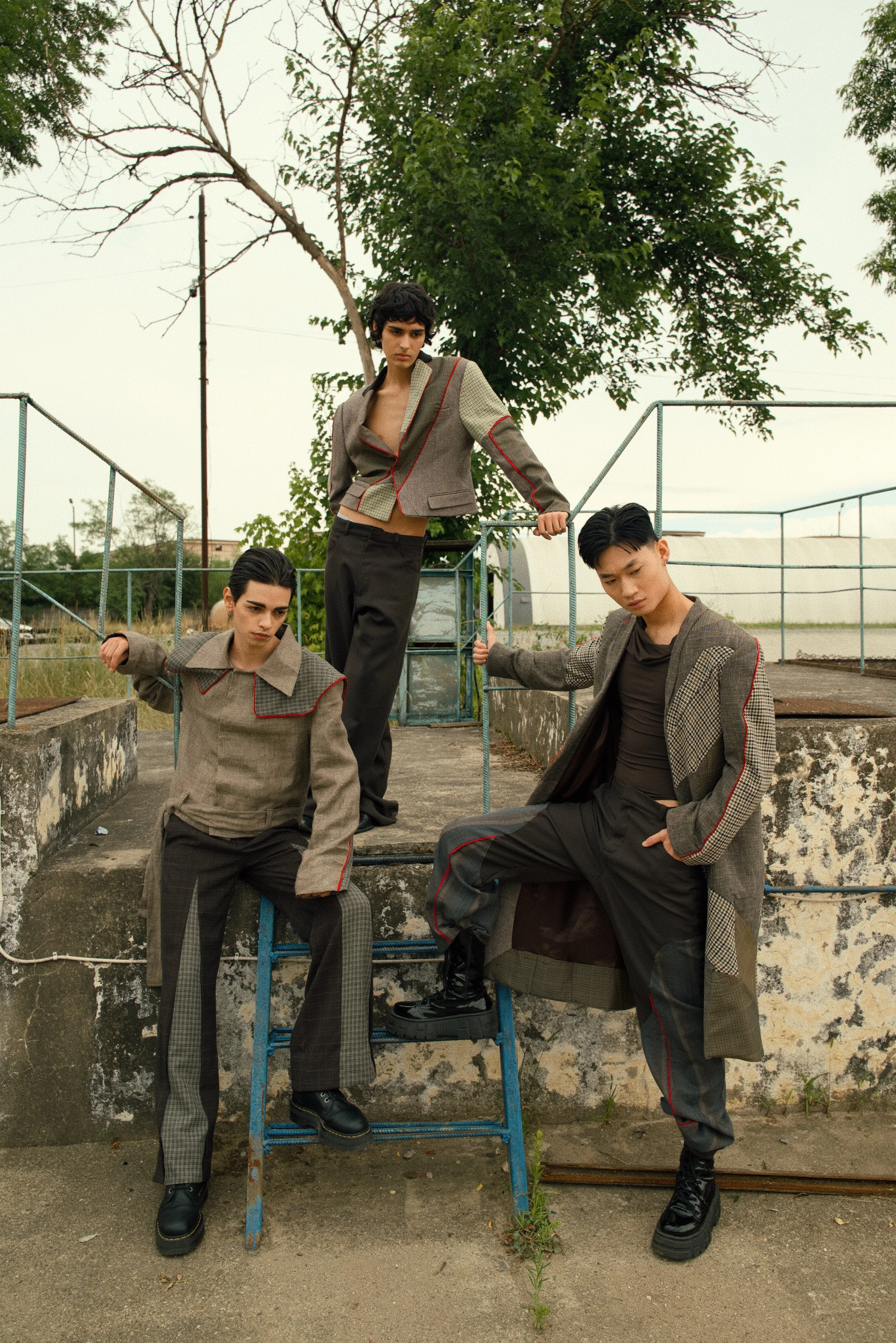 Four models wearing checkered and plaid suits with red accents are posed outdoors on concrete steps and a rusted blue ladder, with trees and an overcast sky in the background.