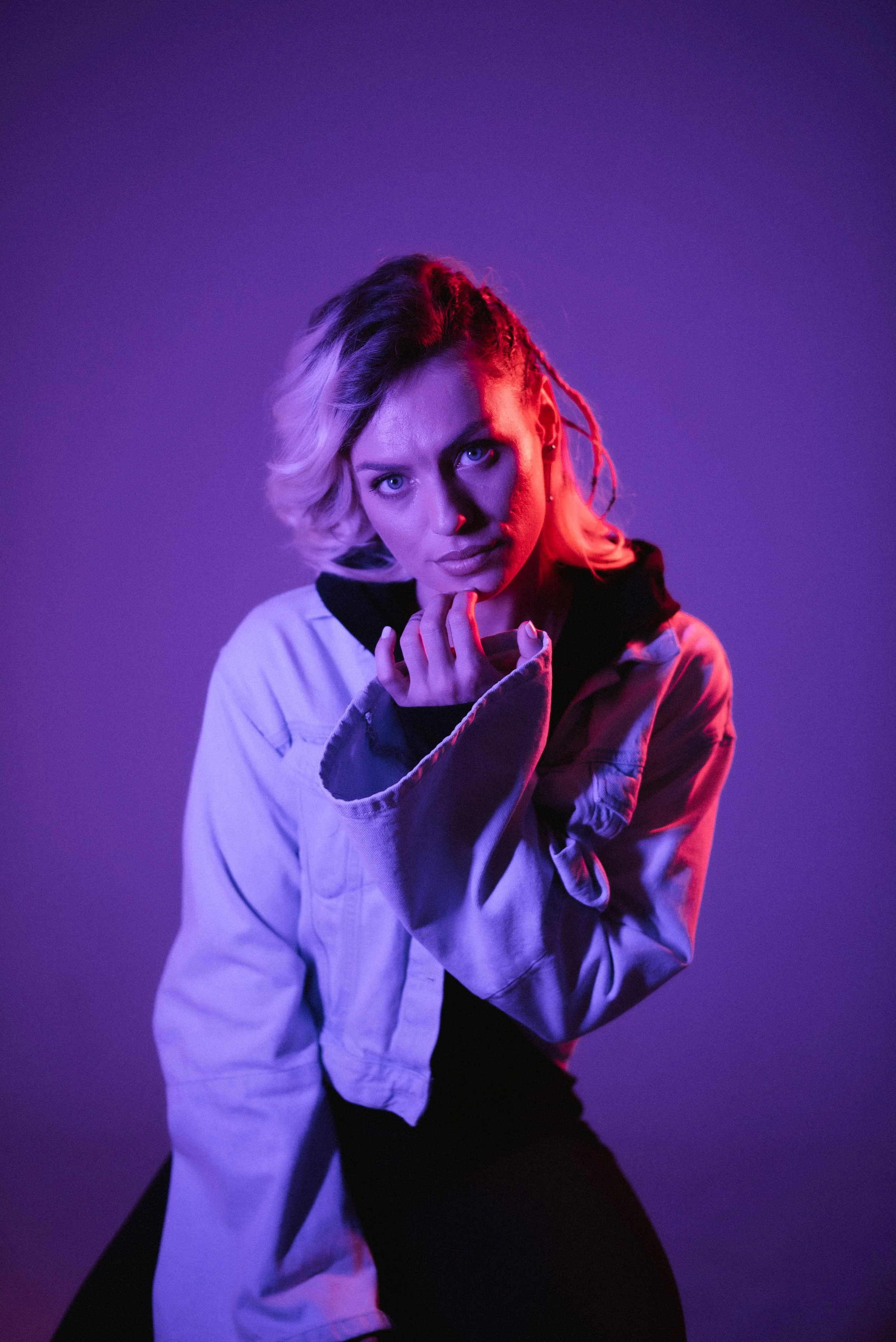 A woman with blonde, wavy hair and light skin poses with her hand near her chin, illuminated with purple and pink lighting.