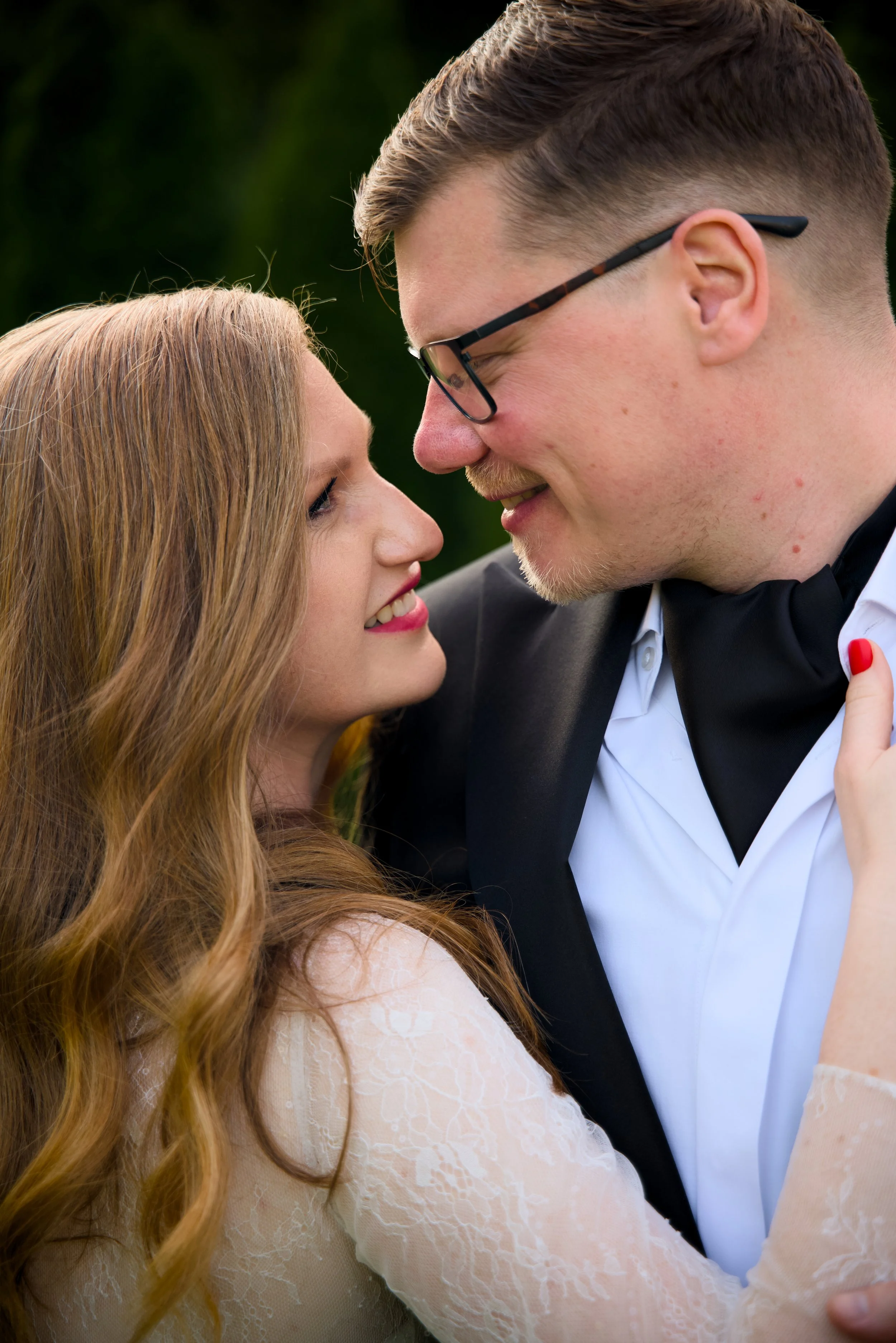A couple with close faces gazing at each other, outdoors, the woman has long red hair and the man has short brown hair and glasses.