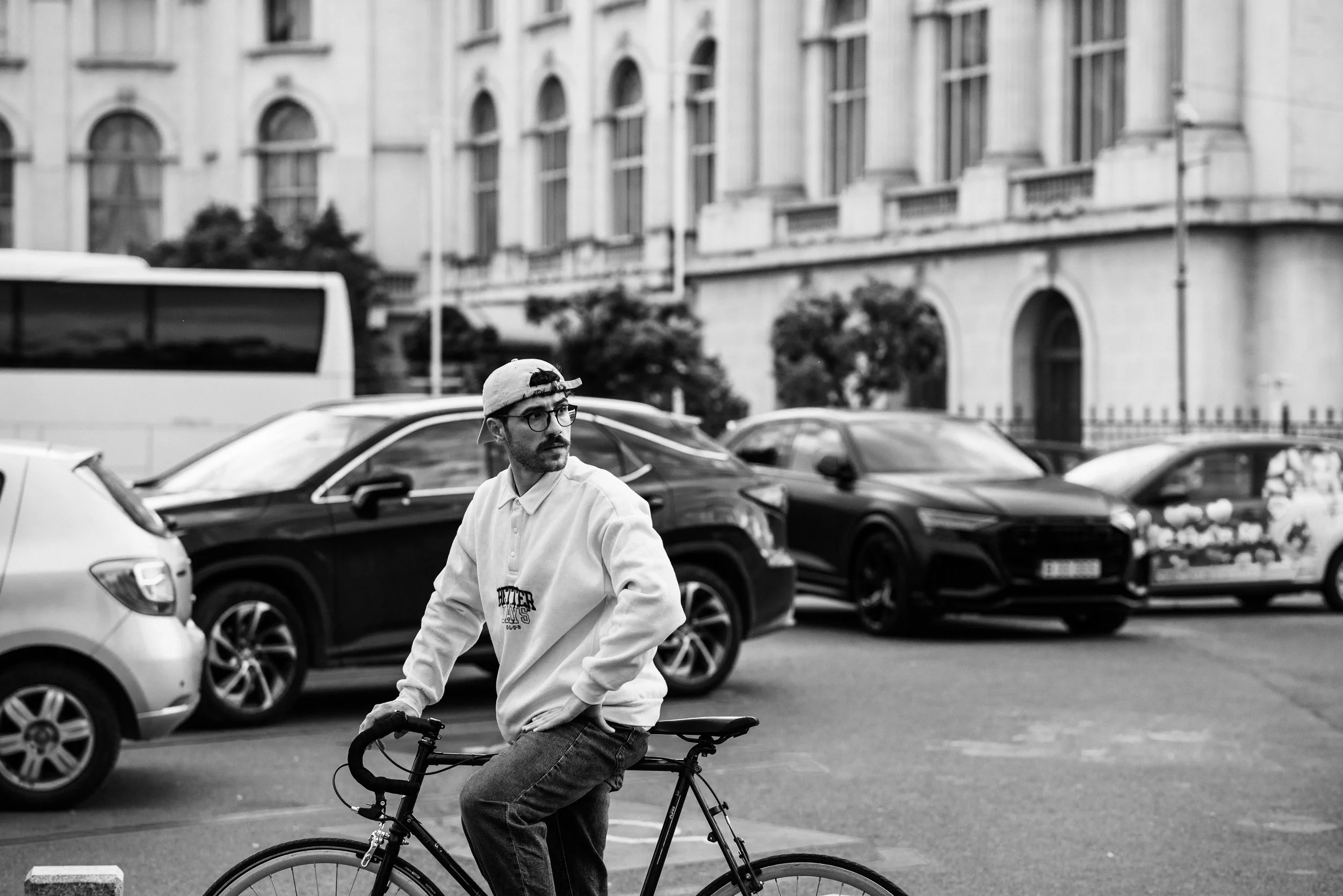 A man with glasses, a beard, and a cap, sitting on a bicycle in a parking lot with cars and a large building in the background.