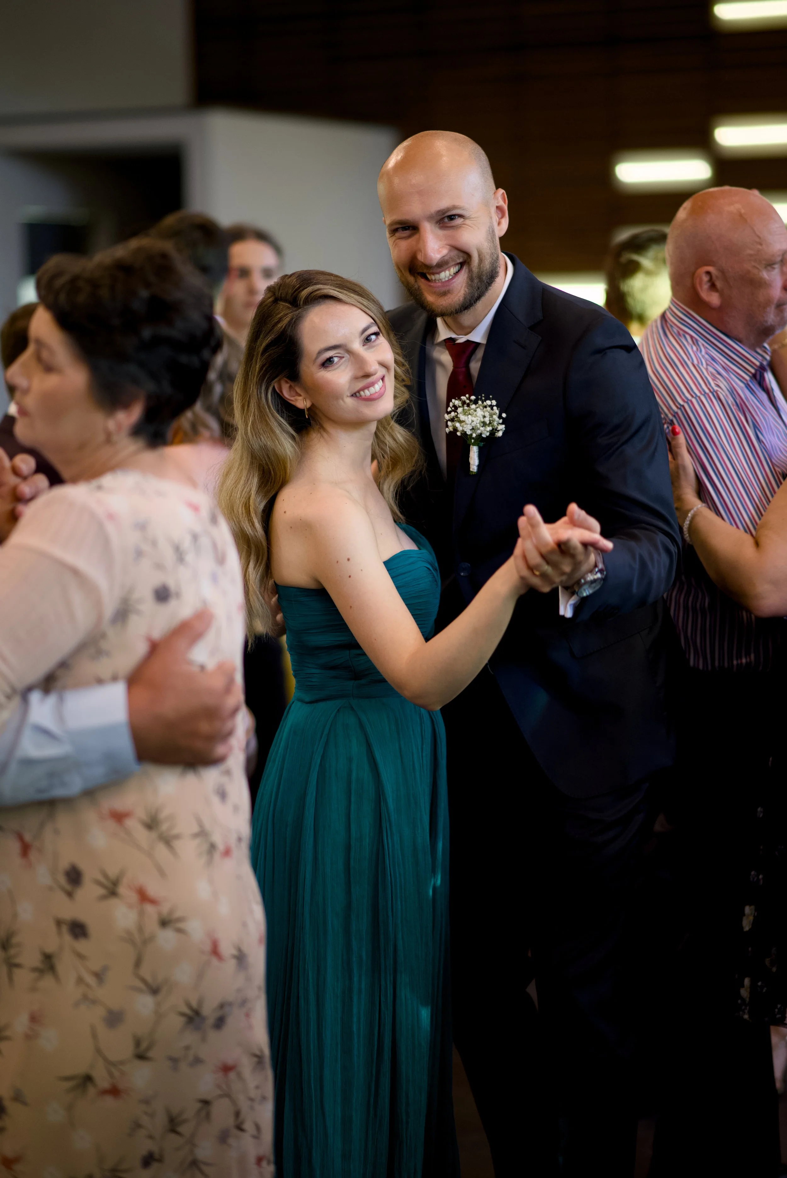 A couple dances together at a social event, with the woman wearing a teal strapless dress and the man in a dark suit with a boutonniere. They are smiling and holding hands, surrounded by other people dancing and chatting indoors.