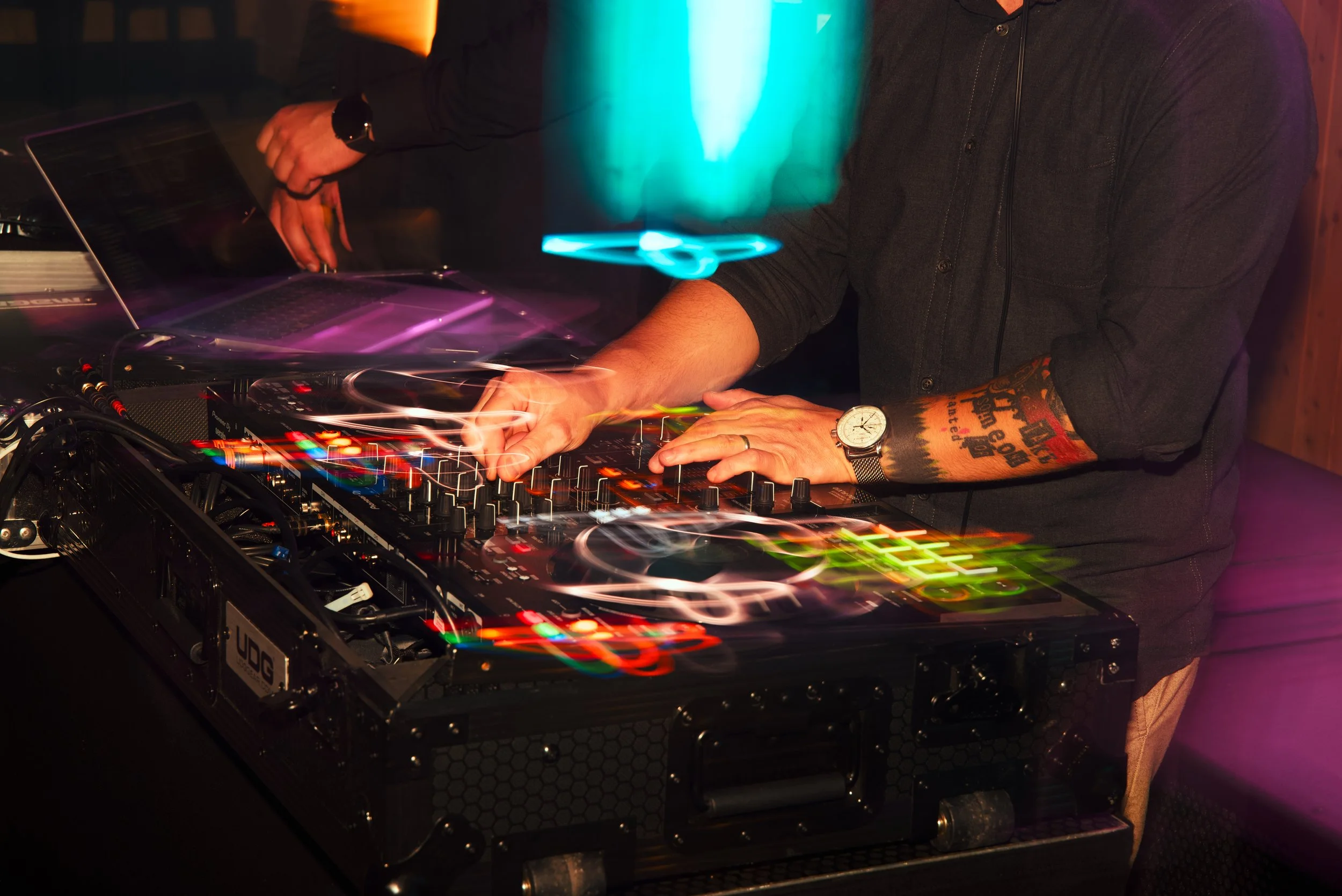 DJ wearing a black shirt and a watch, using a DJ mixer with colorful lights and motion blur, in a dark setting.