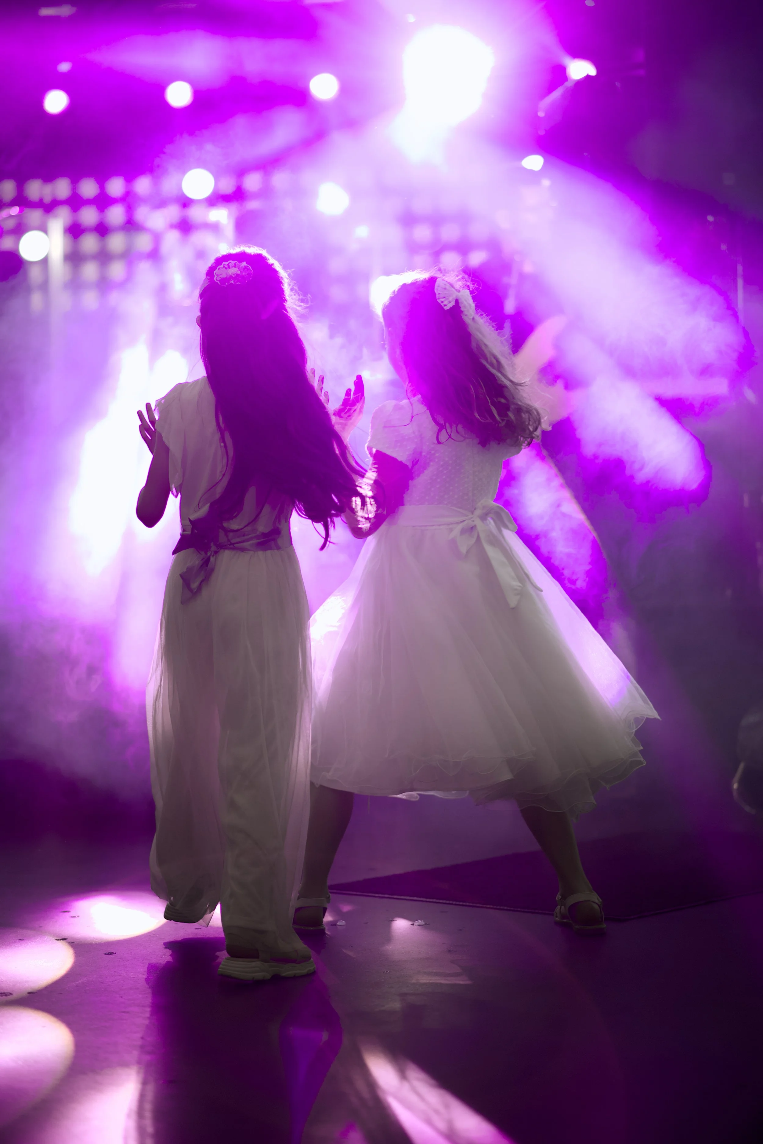 Two young girls dressed in white ballet costumes dancing on a stage with purple and pink lighting.
