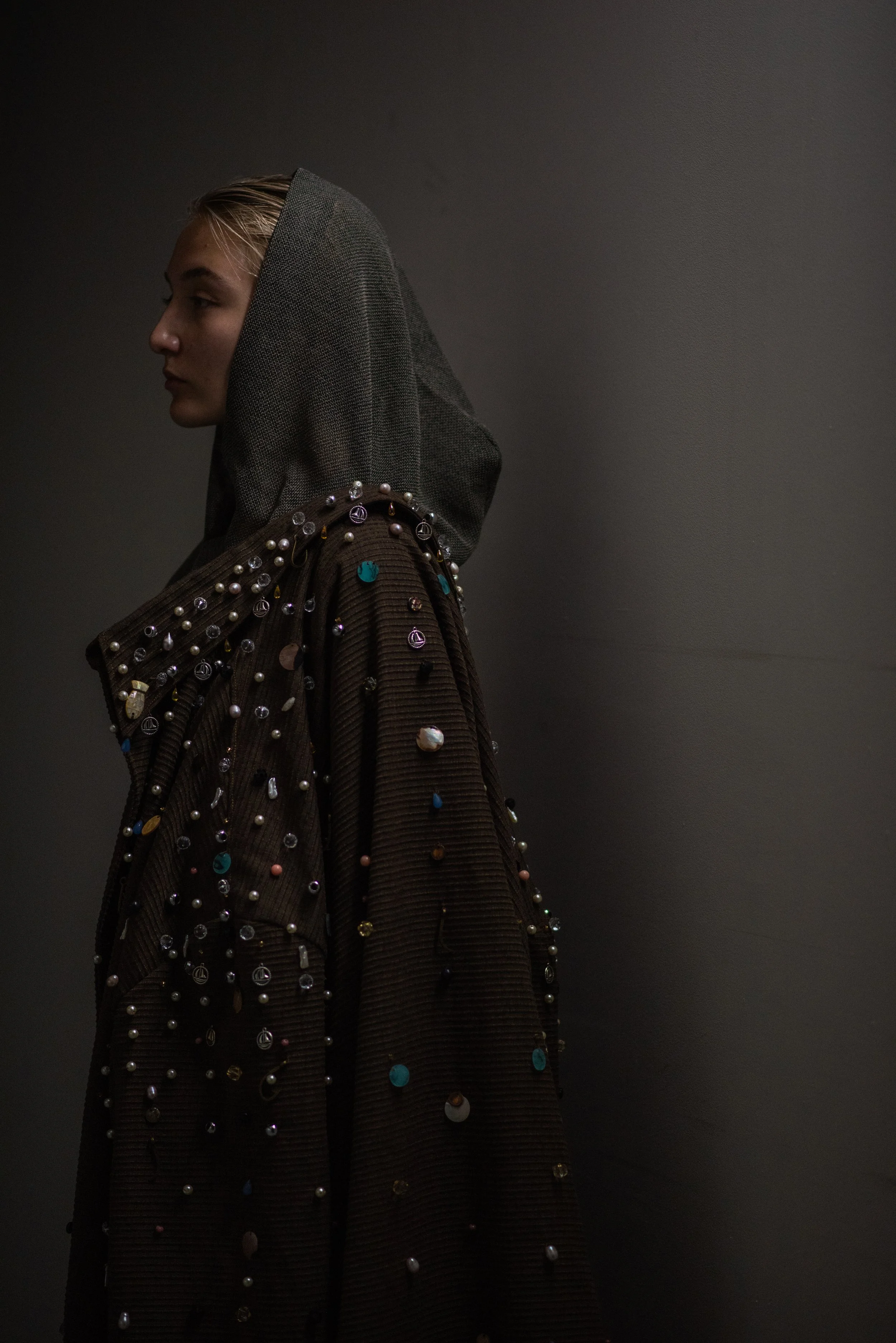 Profile of a woman wearing a dark hooded garment decorated with various beads and embellishments, against a plain dark background.