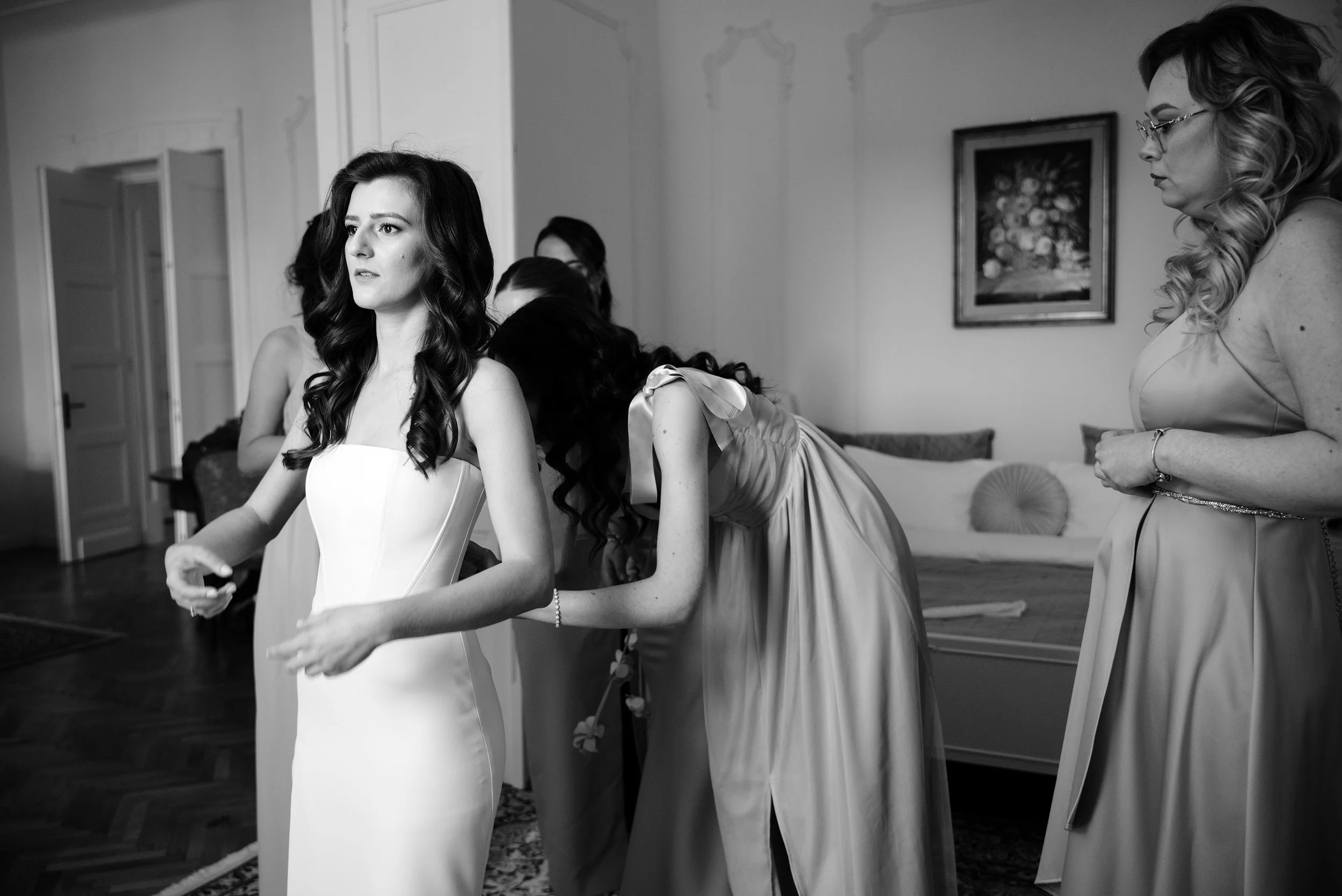 A bride in a strapless white gown getting ready, surrounded by her bridesmaids in a room, some adjusting her dress, in black and white.