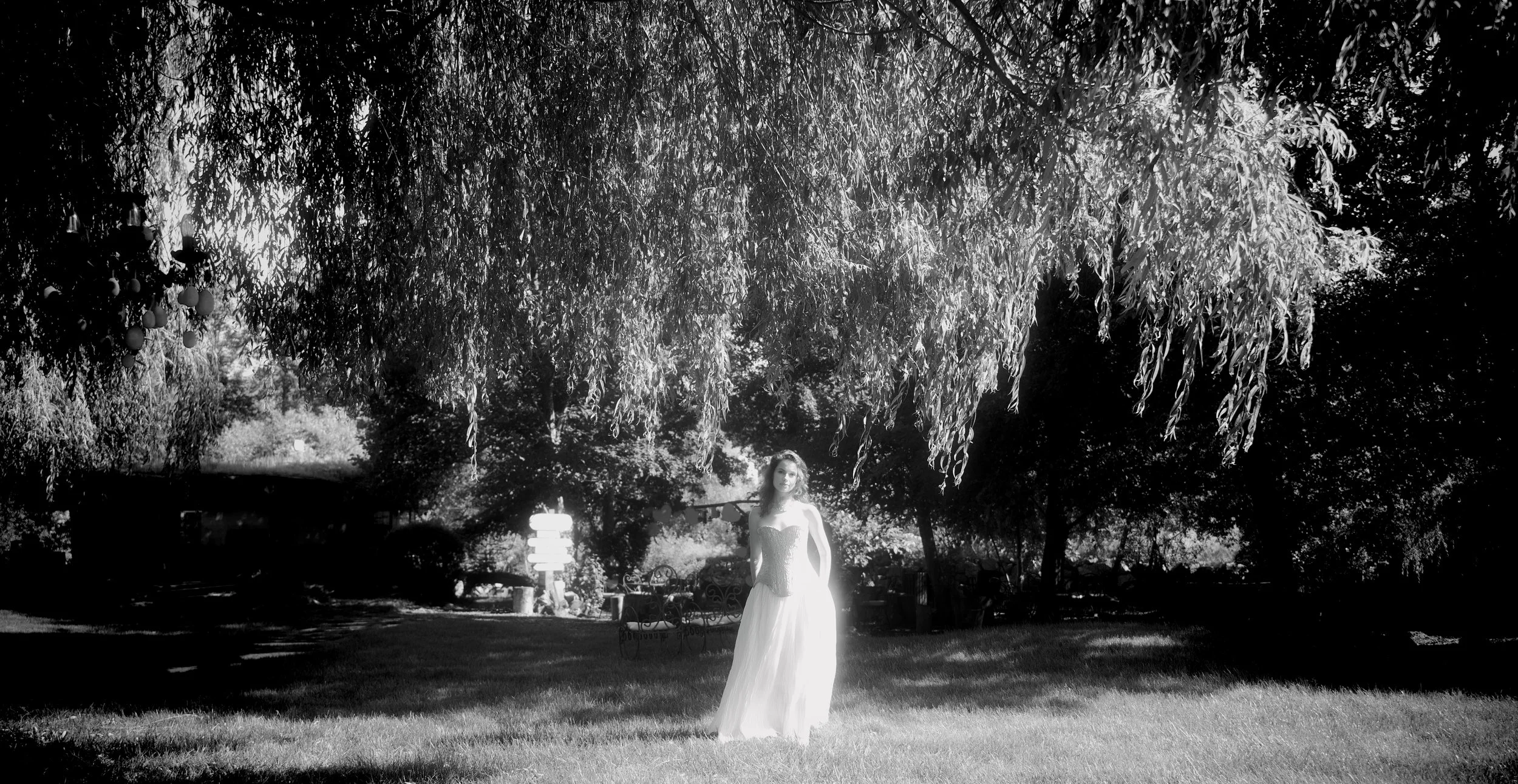 A woman in a strapless dress standing under a large tree in a garden, with picnic tables and benches in the background.