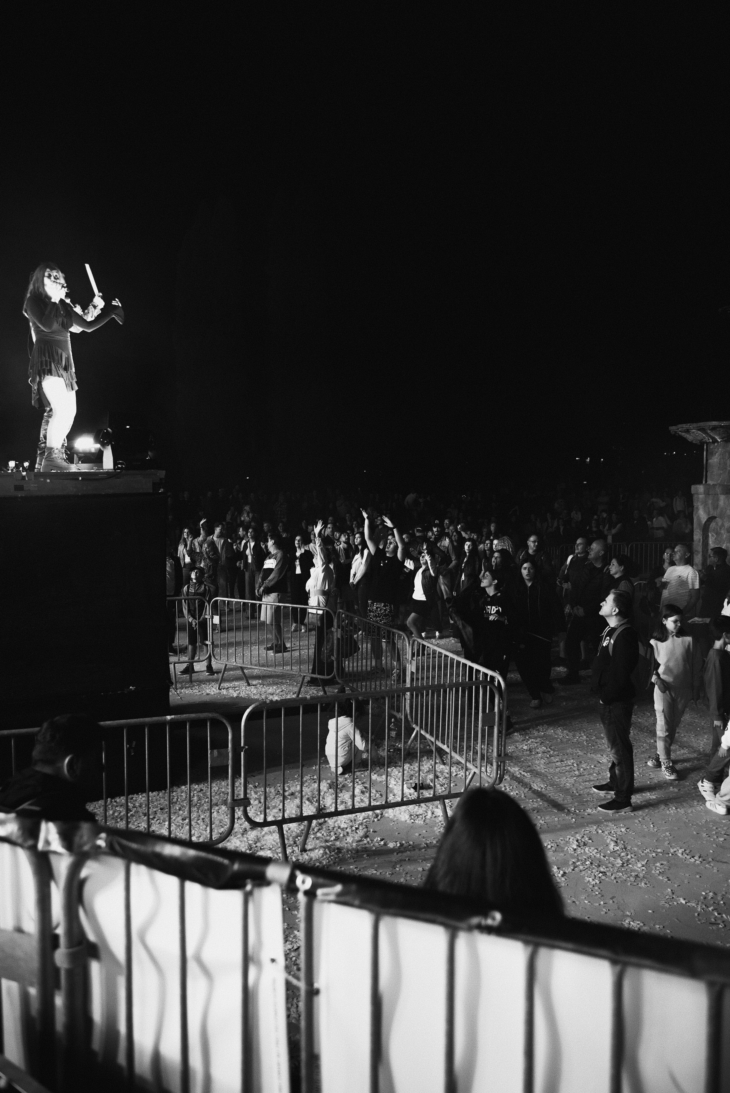 Black and white photo of a concert scene at night with a performer on a stage on the left and a crowd of people dancing and standing in front of the stage.